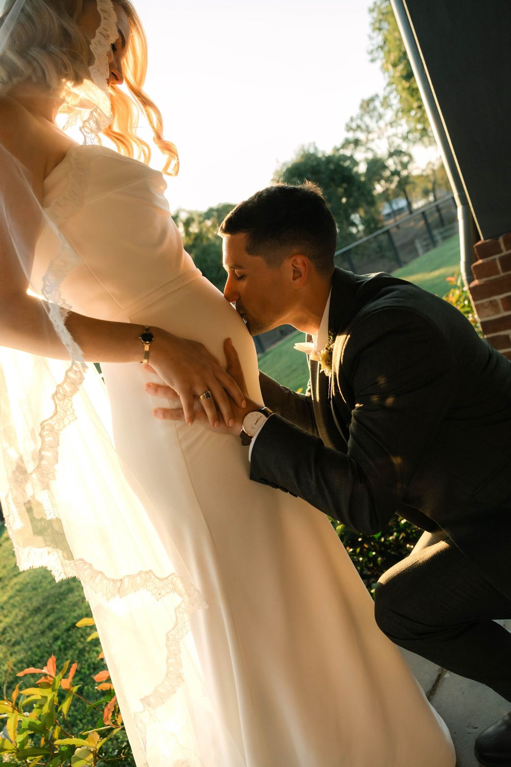 Groom in dark suit kneels to kiss pregnant bride's belly in golden sunset wedding photo.