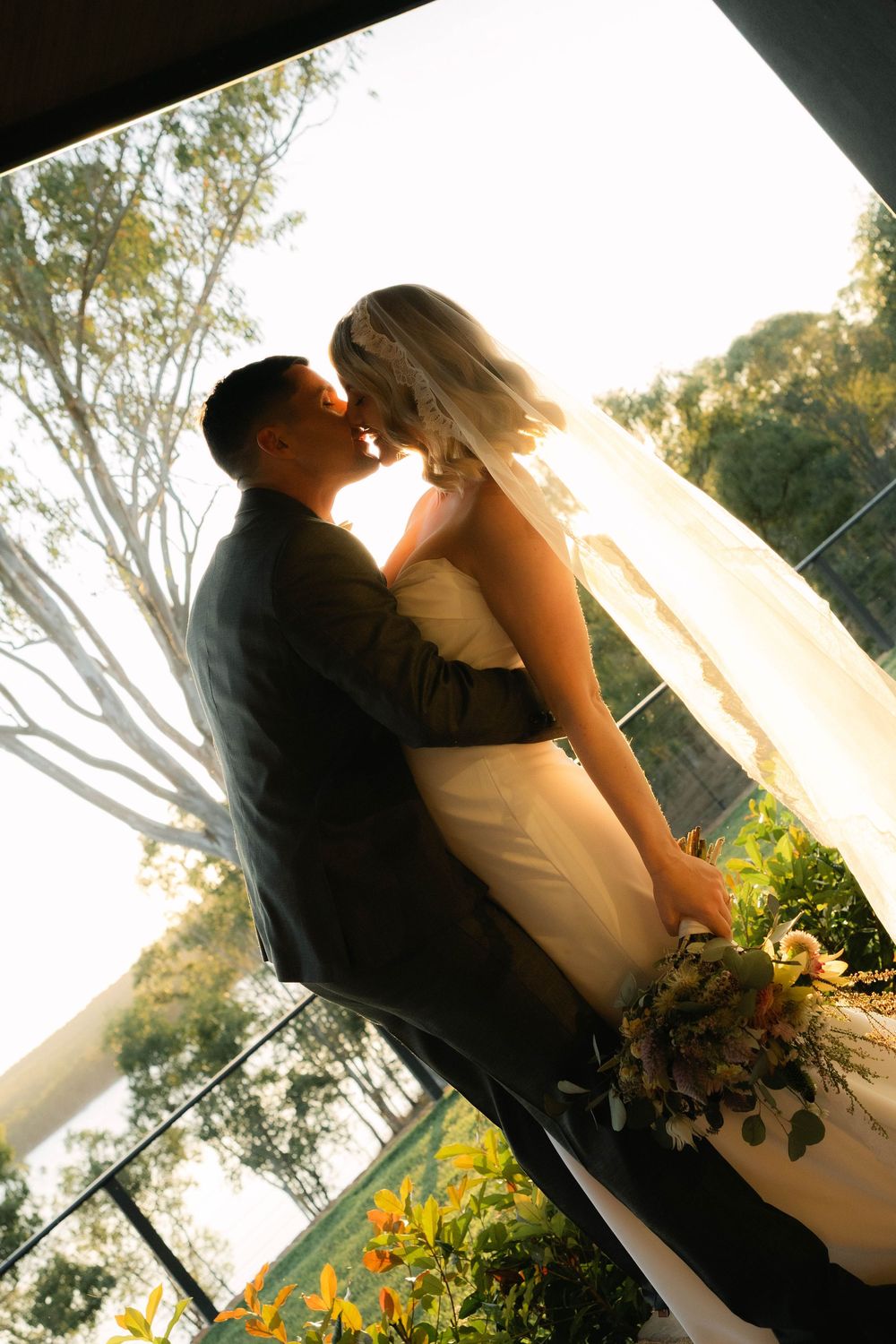 Bride and groom share a romantic kiss at golden hour, with bride holding a bouquet and veil flowing, surrounded by lush greenery.