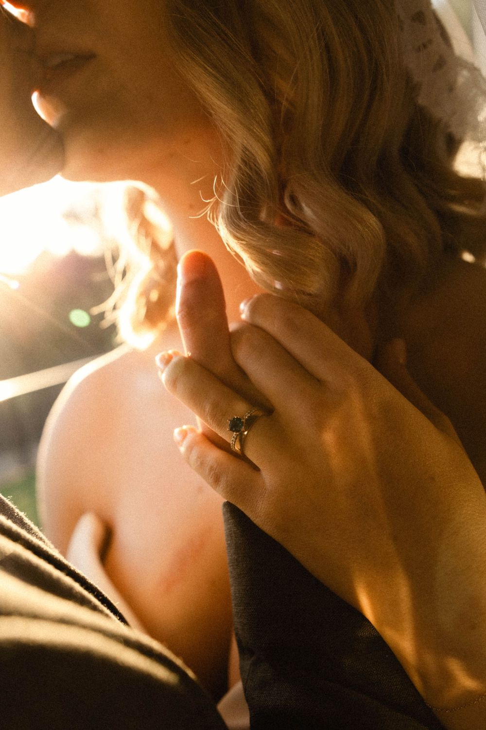 Close-up of bride's engagement ring with dark gemstone as groom tenderly touches her face at sunset.
