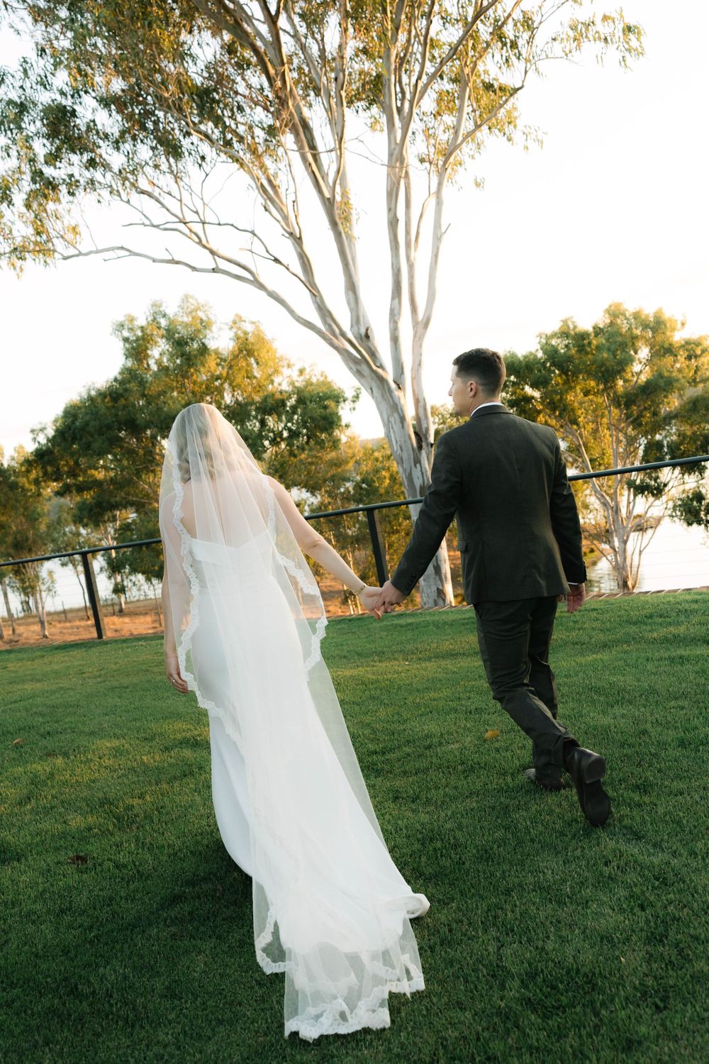 Bride and groom walking hand in hand across green lawn toward trees, bride wearing white gown with long veil.