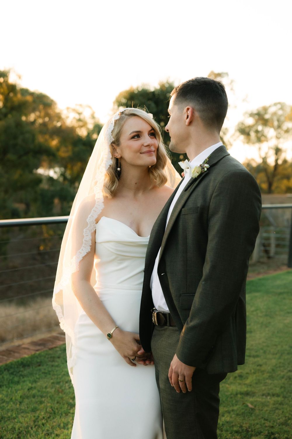 Bride in white gown and veil gazes at groom in olive suit during golden hour outdoor wedding portrait.
