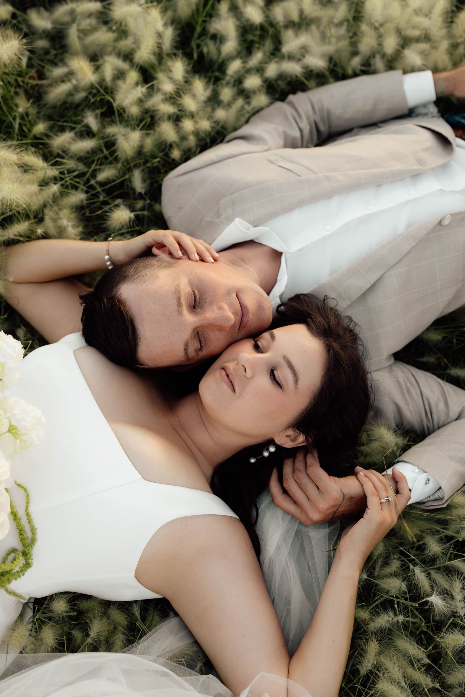 Bride and groom lying in a field of tall grass, eyes closed, sharing a peaceful romantic moment on their wedding day.