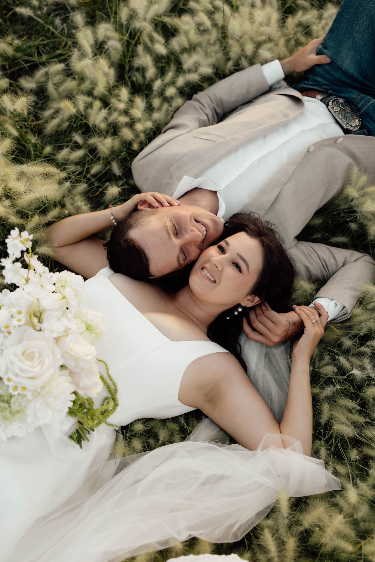 A smiling bride and groom lying in a field of tall grass, with a white rose bouquet nearby.