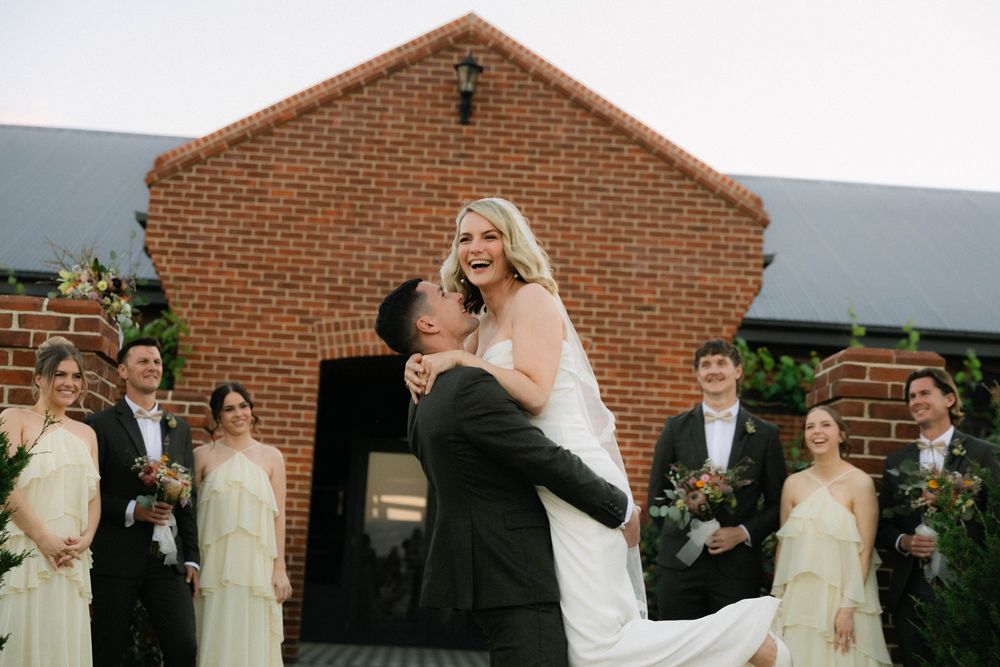Groom lifting bride in white dress outside brick church with wedding party.