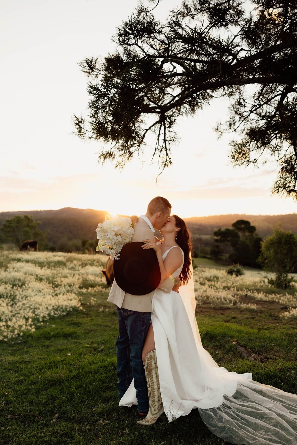 Bride and groom kissing at golden sunset in a field of white wildflowers with rolling hills in the background.