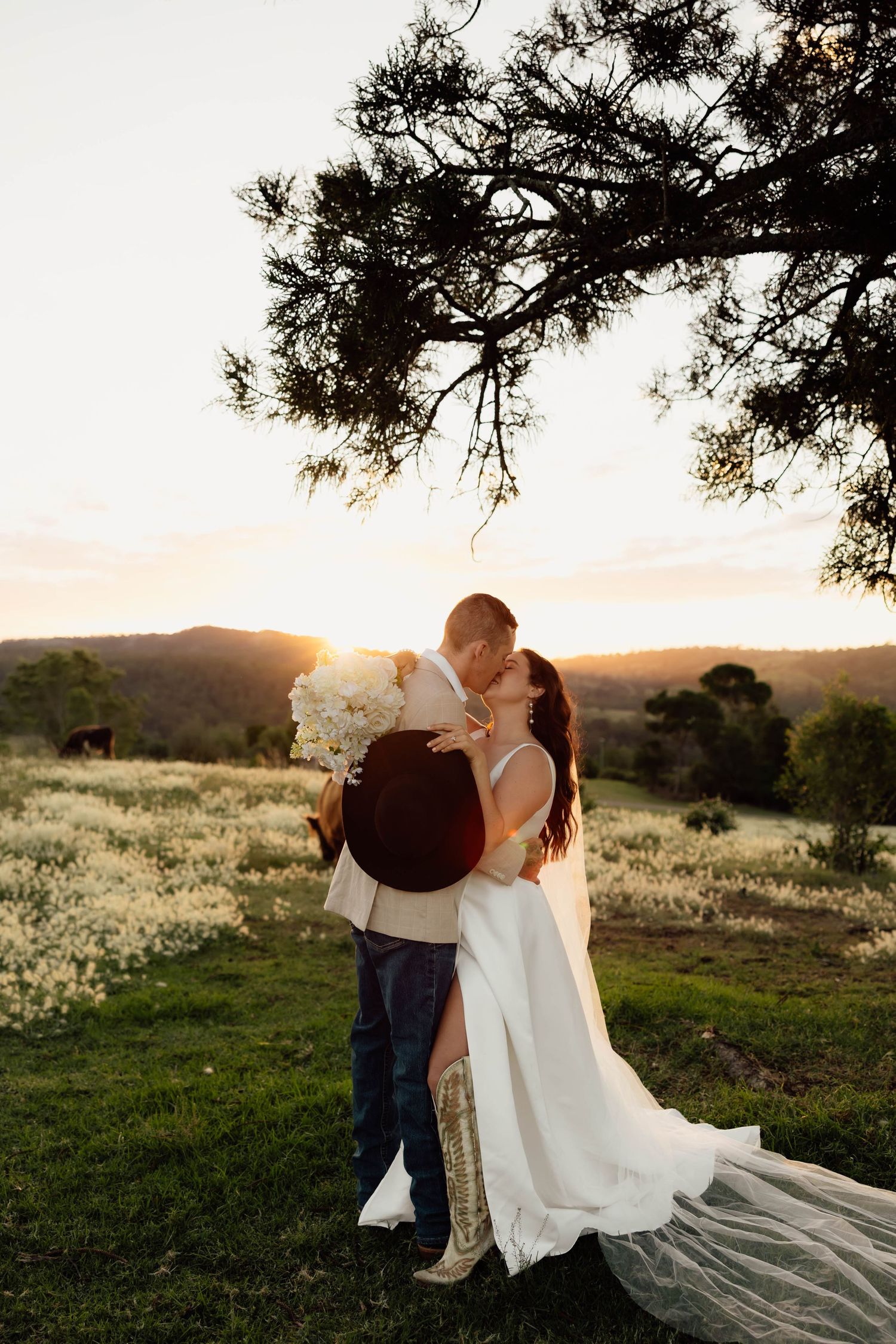 Bride and groom kissing at golden sunset in a field of white wildflowers with rolling hills in the background.