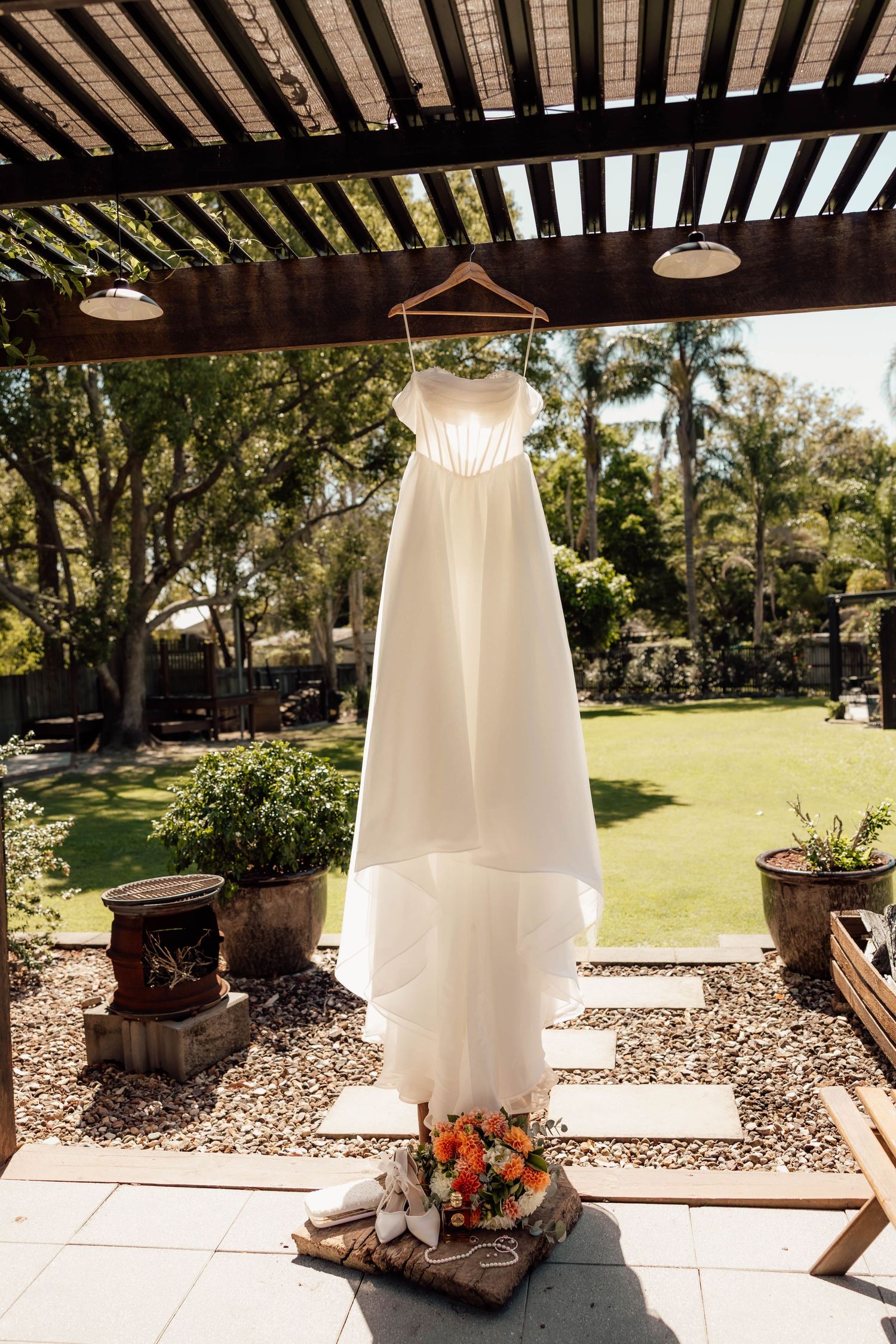 White wedding dress hanging on a wooden pergola outdoors, with a bridal bouquet of orange flowers below.