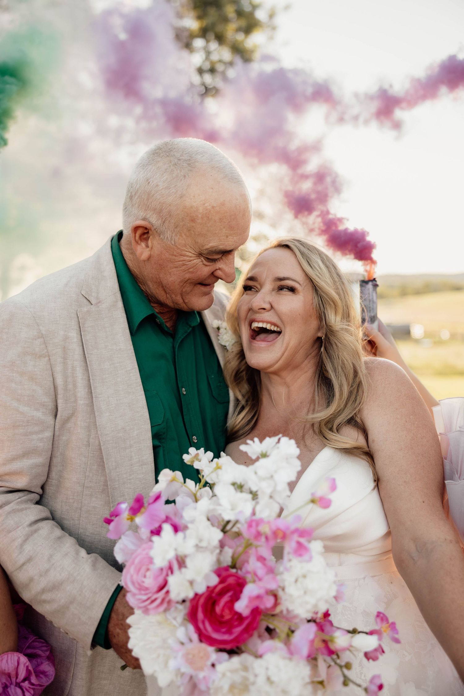 Joyful bride laughing with older man in green shirt at outdoor wedding with colorful smoke bombs and pink bouquet.