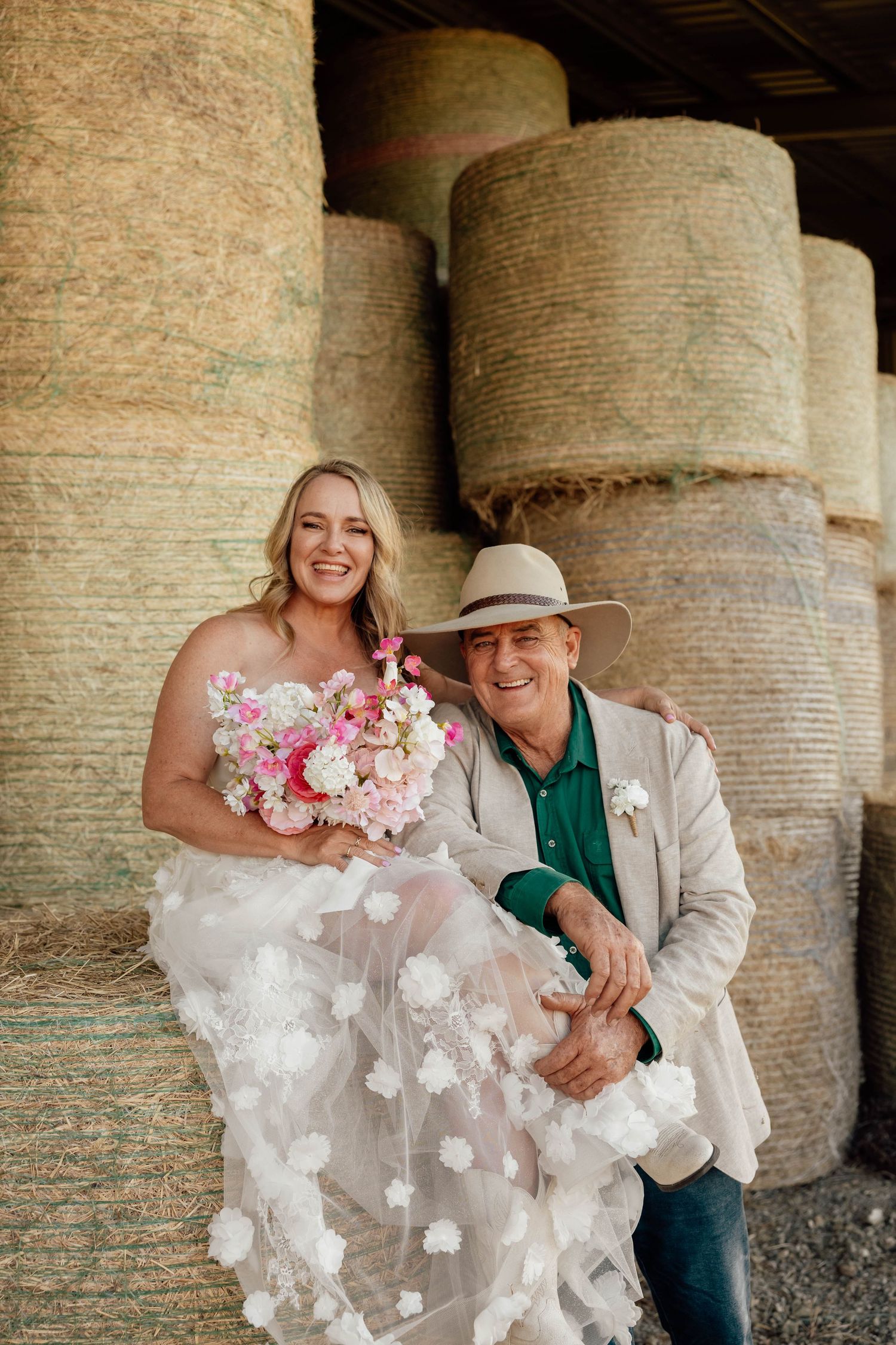 Bride in floral wedding gown holding pink bouquet with man in cowboy hat beside hay bales at rustic barn wedding.