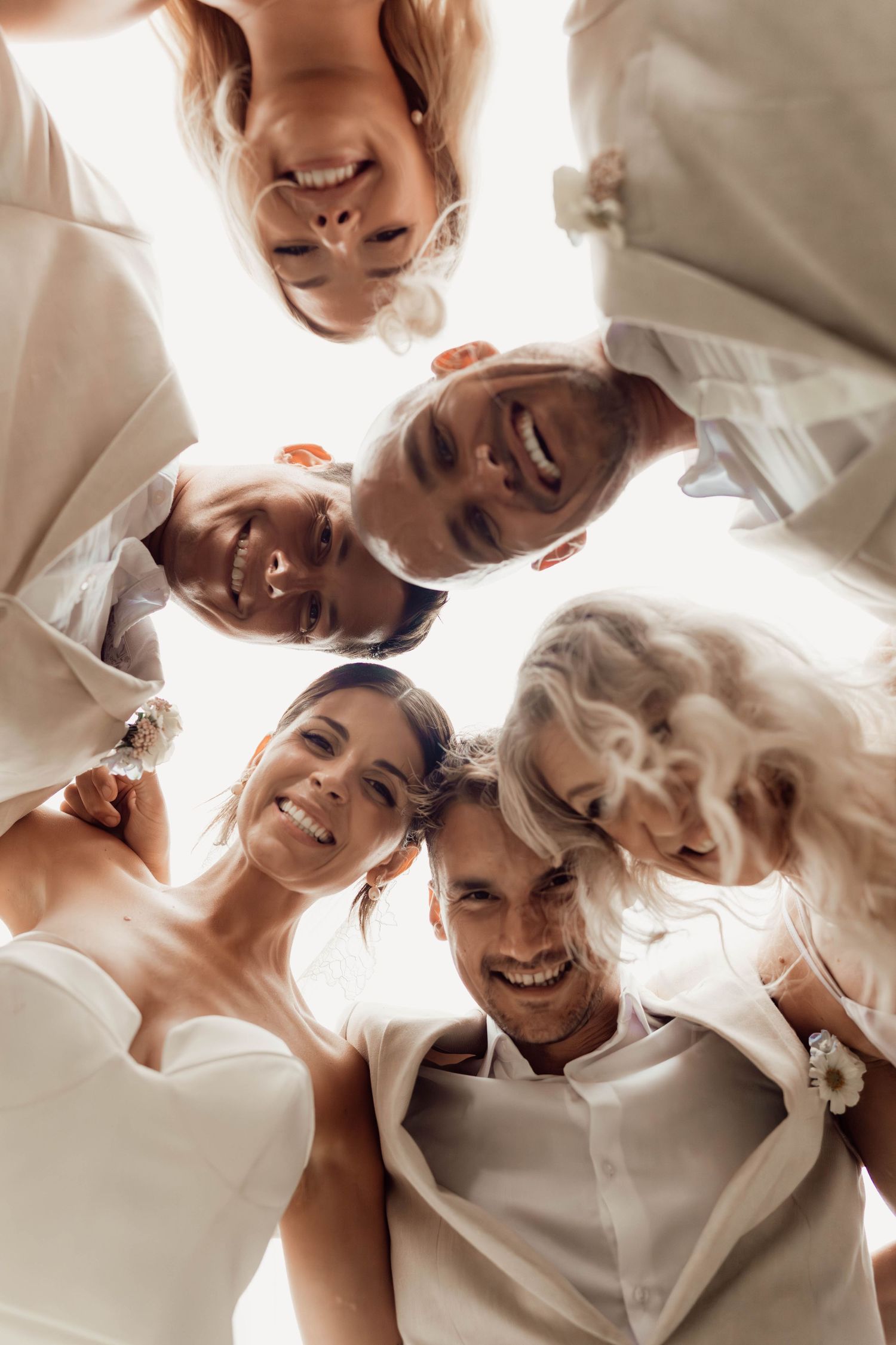 Happy wedding party huddles together smiling in a circle viewed from below in white attire.