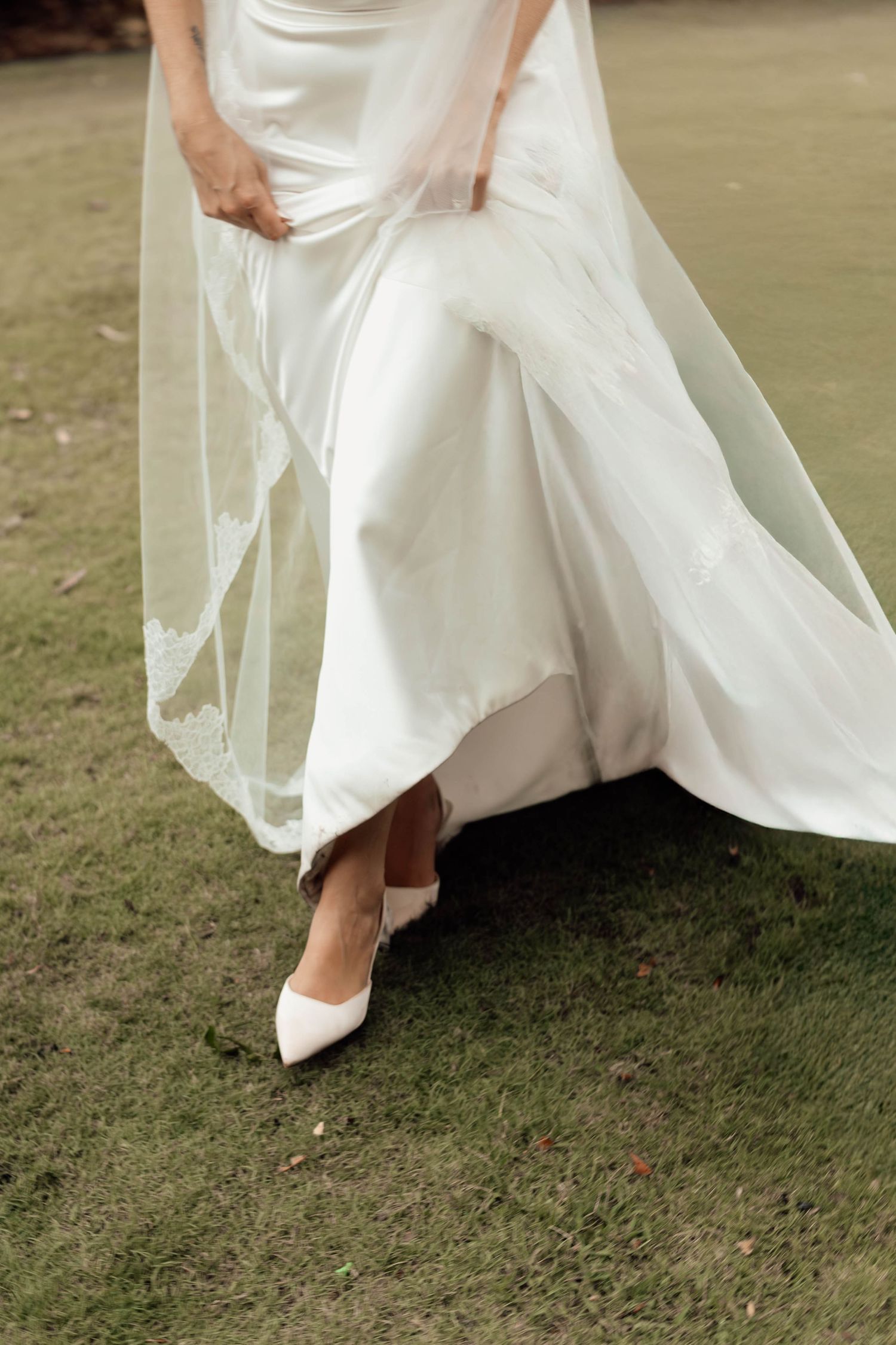 Bride in white satin gown and heels lifting her dress and veil on grass.