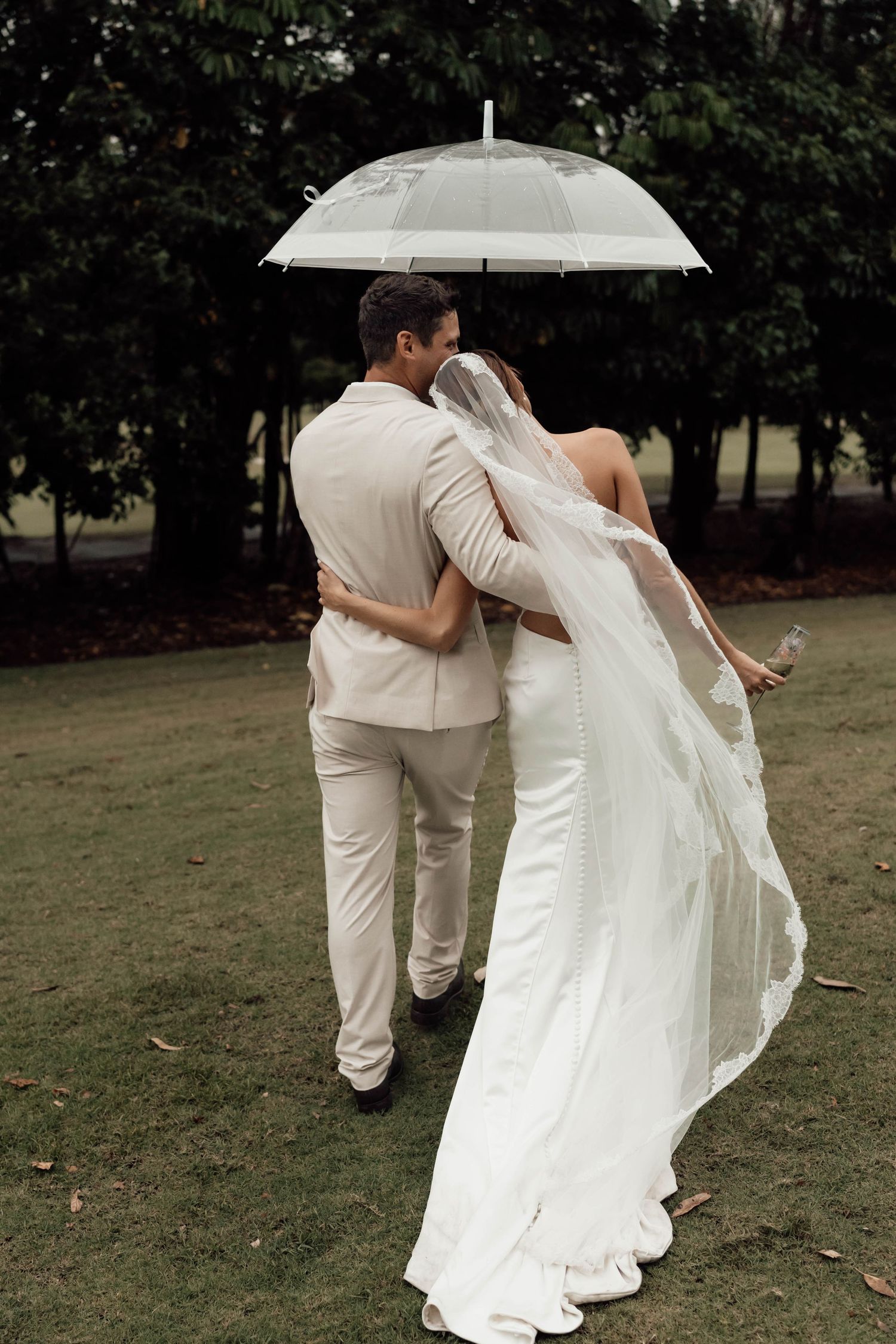 Bride and groom walking under a clear umbrella in a park, bride wearing a flowing white gown with long veil.