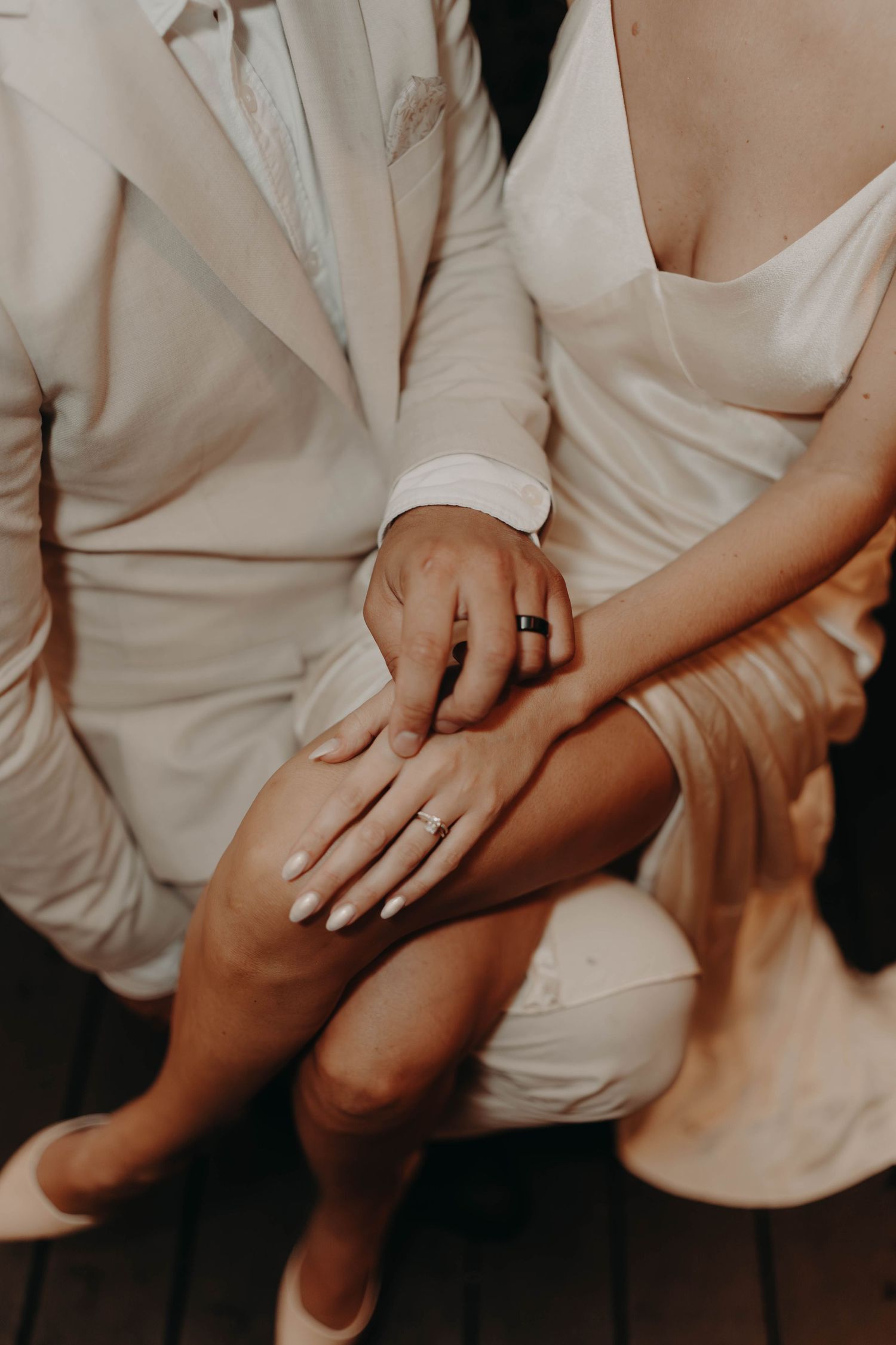 Newlyweds sitting closely together showing off their wedding rings, bride in white dress and groom in white suit.
