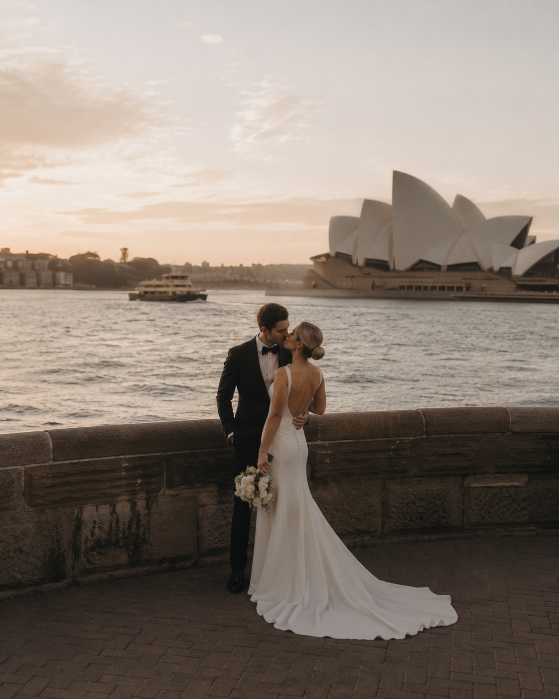 Couple sharing a romantic kiss in wedding attire with Sydney Opera House at sunset backdrop.