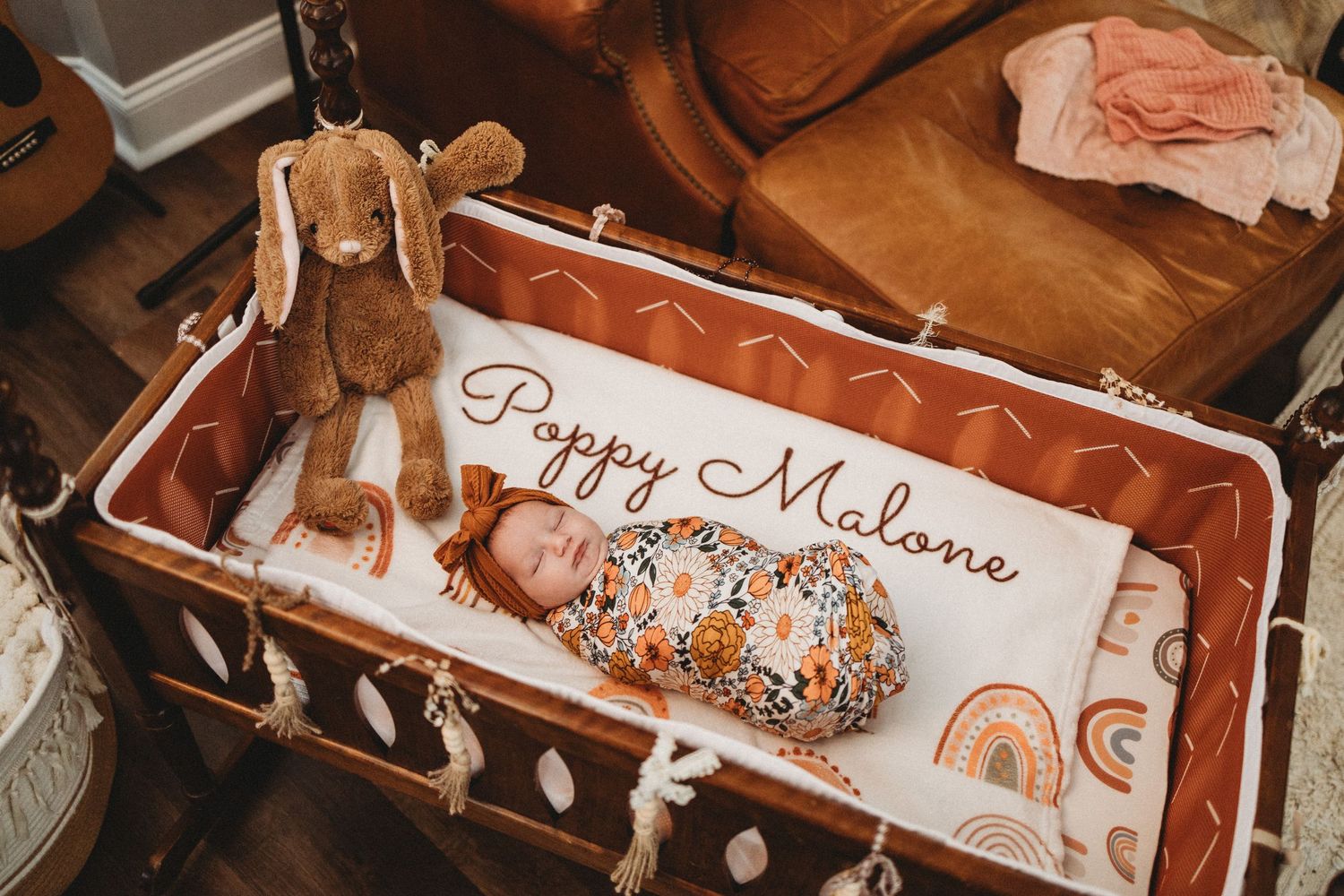 Newborn baby swaddled in floral wrap sleeping in a wicker bassinet with a stuffed bunny and Poppy Malone name sign.