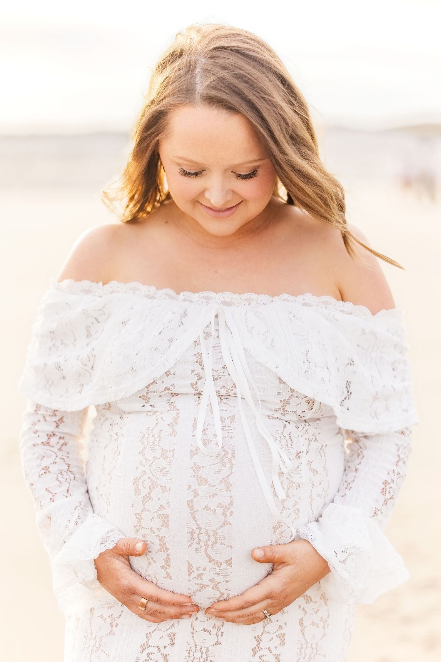 Pregnant woman in white off-shoulder lace dress cradling baby bump on beach, smiling down softly in golden light.
