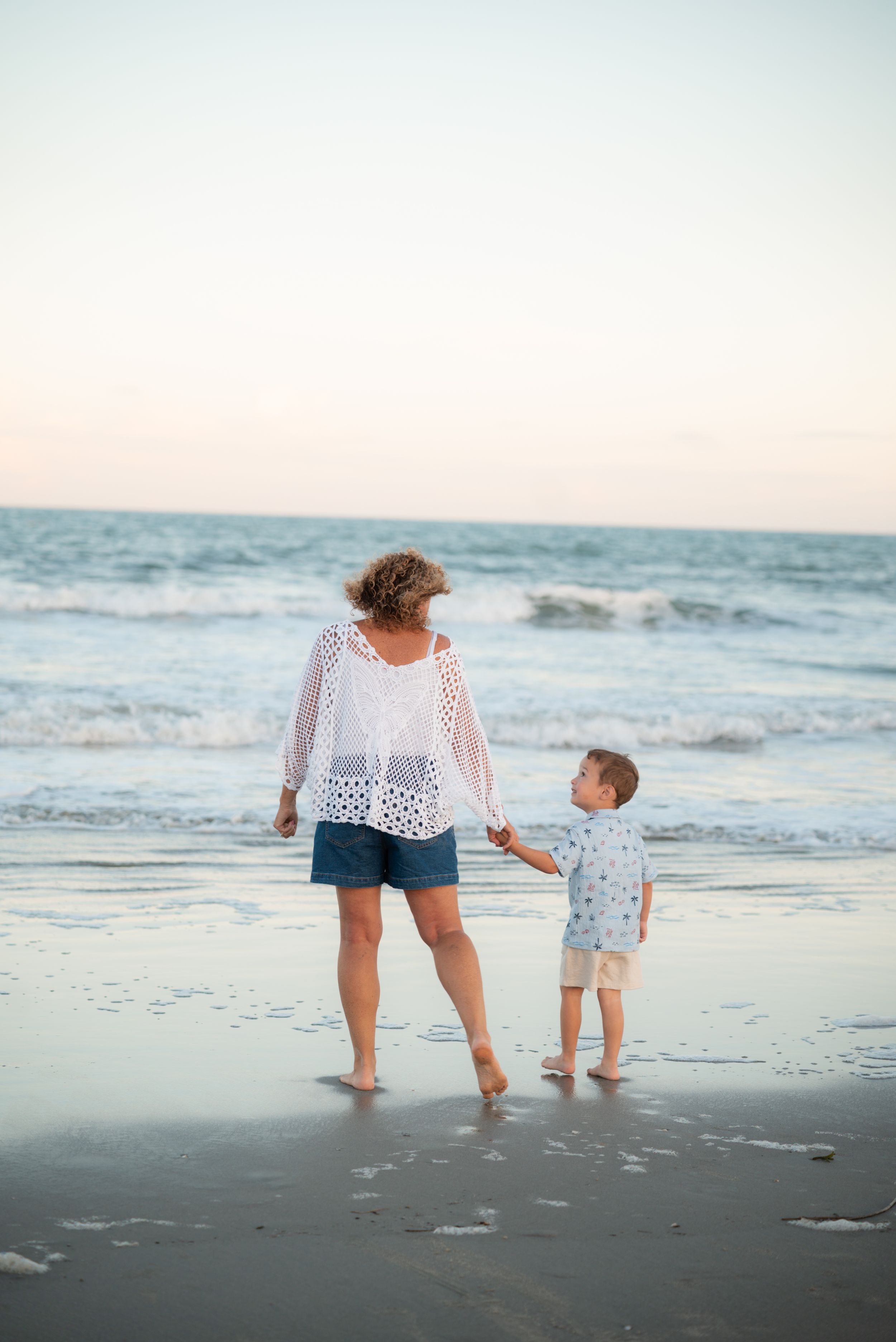A woman and young boy hold hands while standing barefoot on a sandy beach near the ocean at dusk.