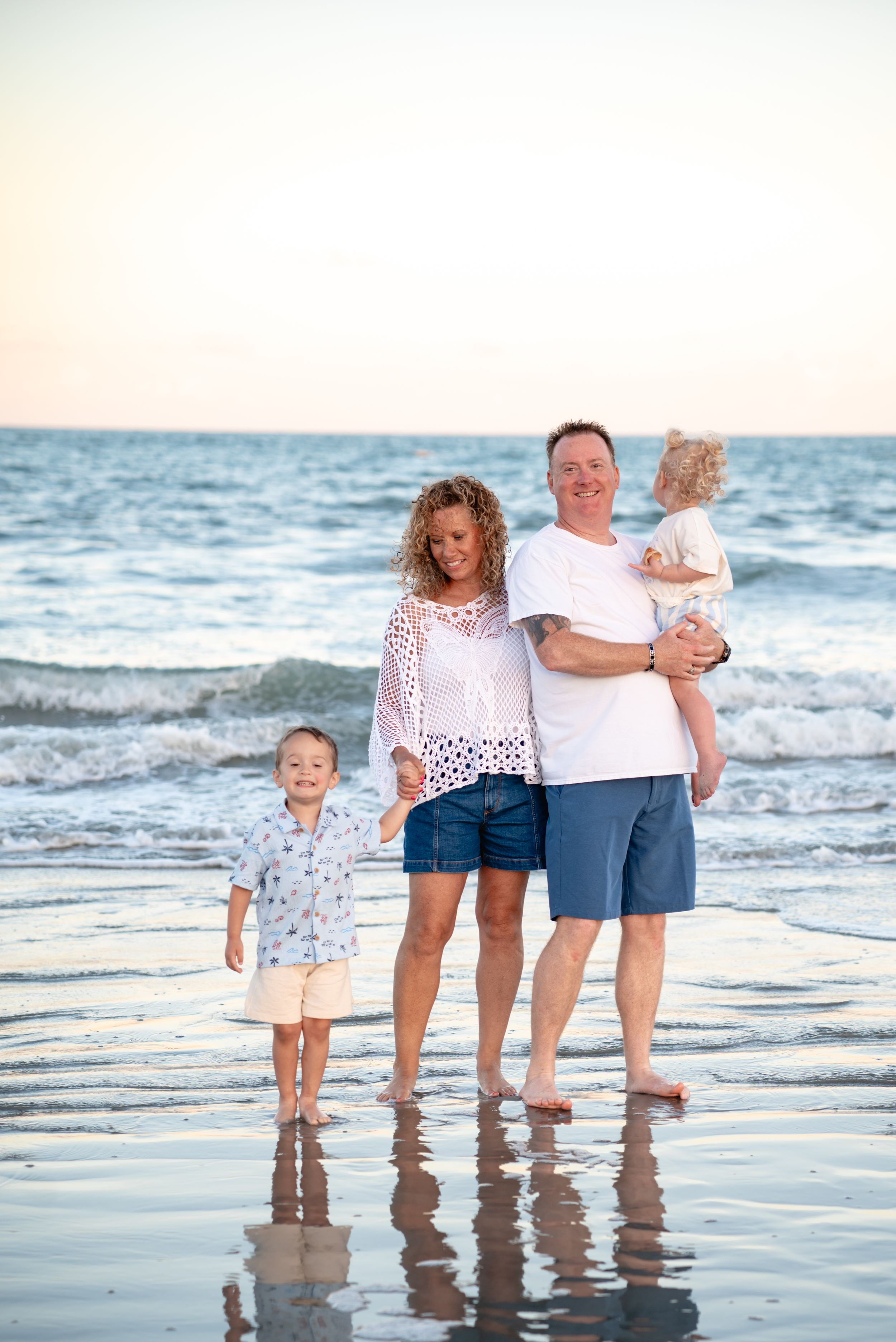 Happy family of four posing on a sandy beach at sunset with ocean waves in the background.