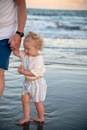 Toddler in striped shorts holds adult's hand while standing in ocean waves at sunset on a sandy beach.
