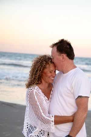 Happy couple embracing and sharing a kiss on a beach at sunset, woman in white crochet top smiling.