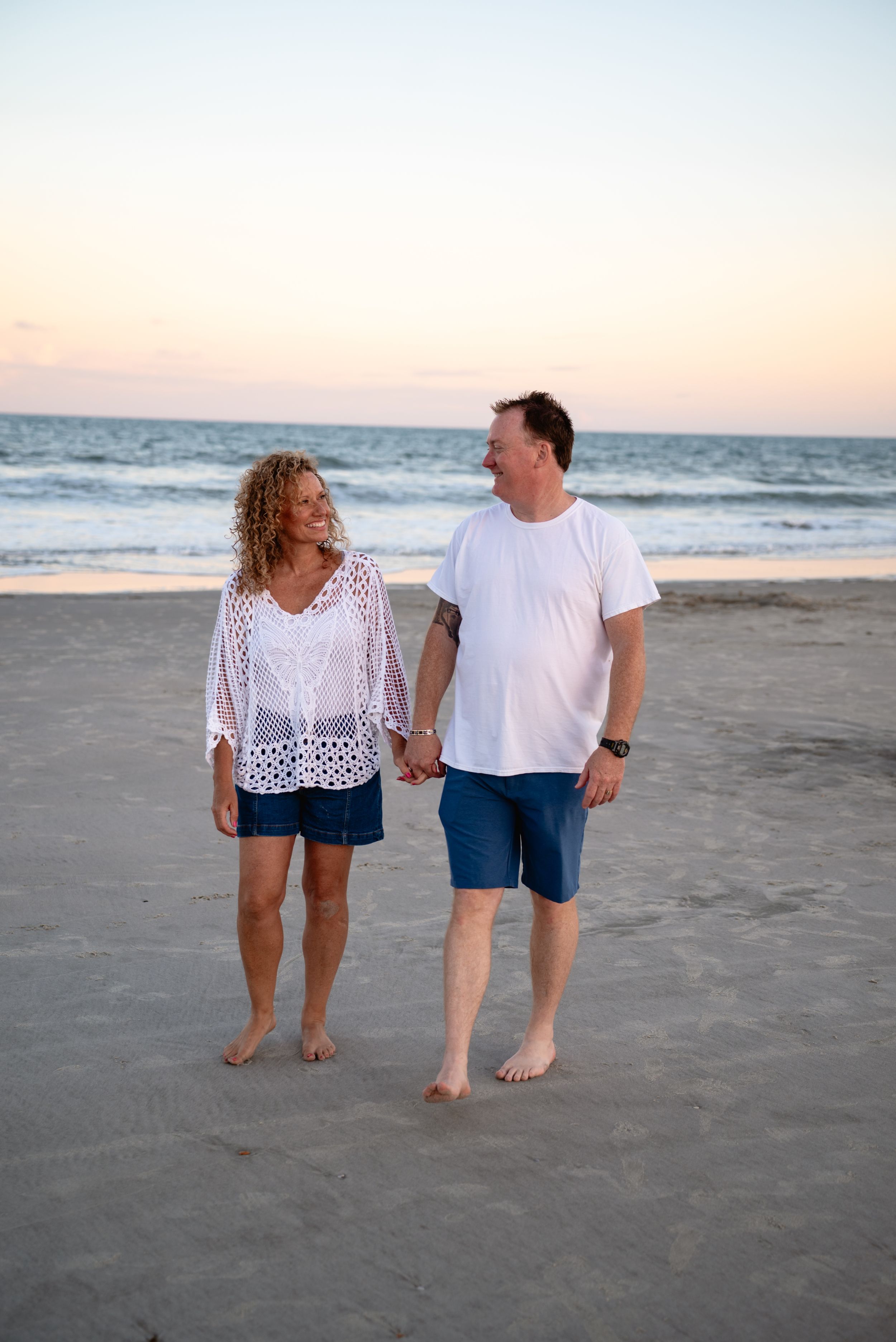 A couple holds hands and smiles while walking barefoot on a sandy beach at sunset.