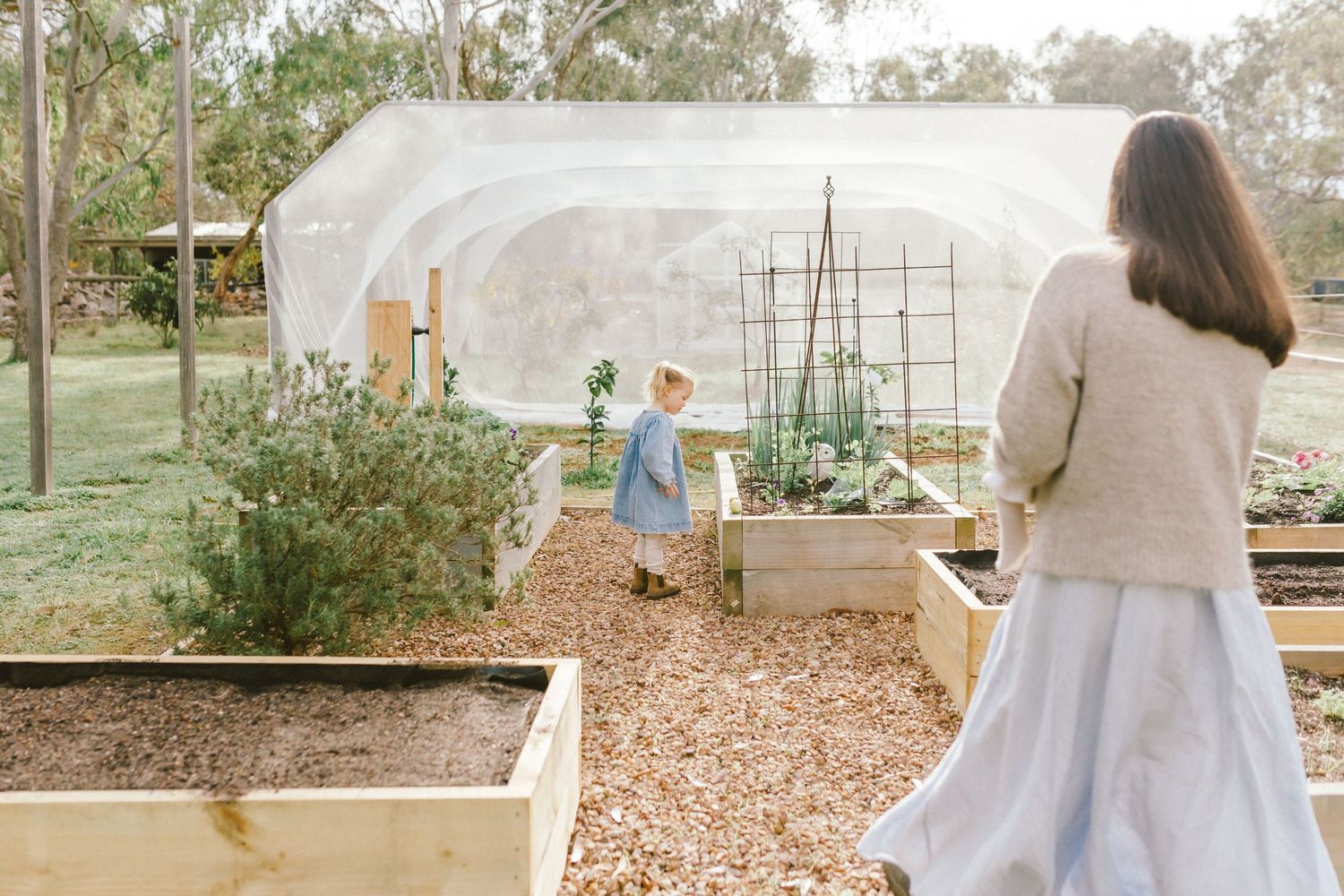 A mother and child explore raised garden beds near a greenhouse tunnel in a lush backyard garden.
