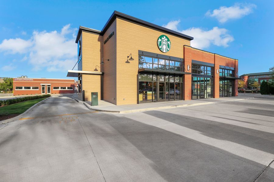 Modern Starbucks coffee shop exterior with large glass windows, drive-through lane, and brick facade under a blue sky.