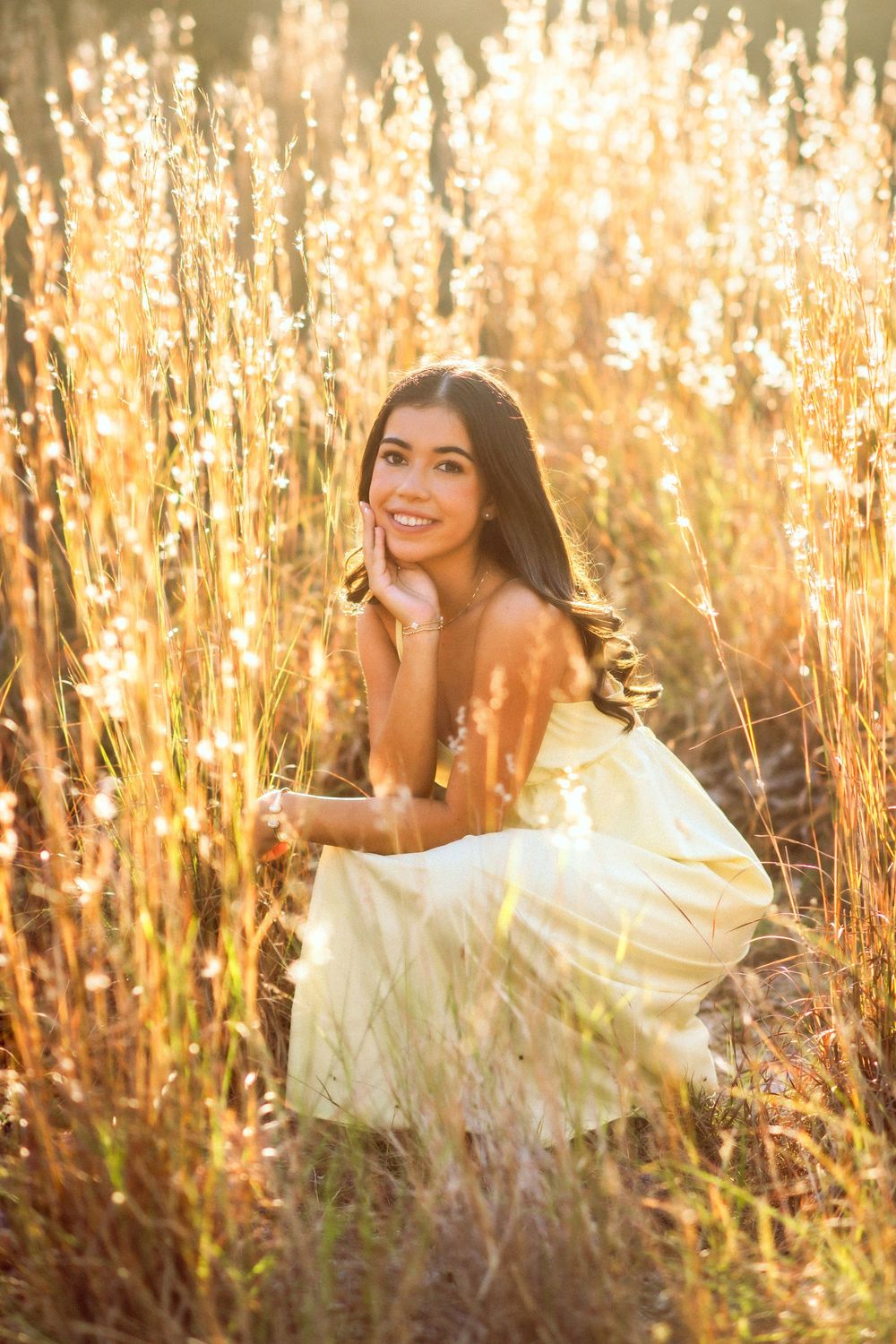 Beautiful young woman crouching in golden tall grass wearing yellow dress at sunset.