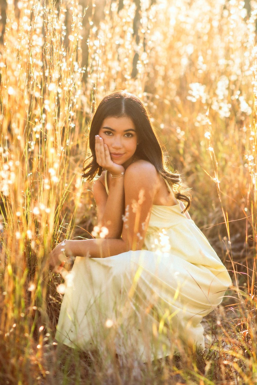 Young brunette woman in yellow dress sitting in glowing golden tall grass at sunset.