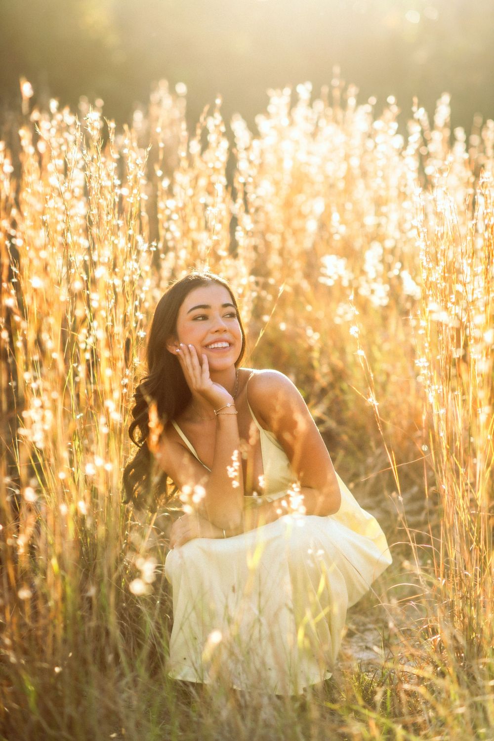 Smiling young woman in yellow dress sitting in glowing sunlit tall grass field.