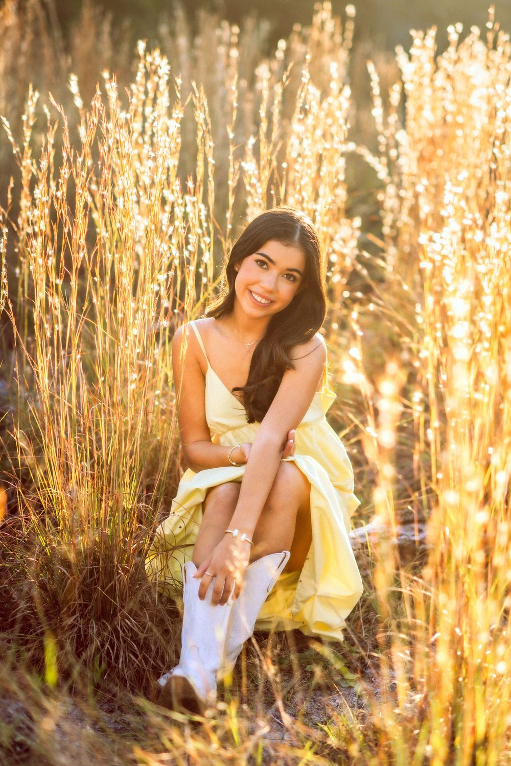Young woman in yellow dress and white cowboy boots sitting in golden sunlit grass.