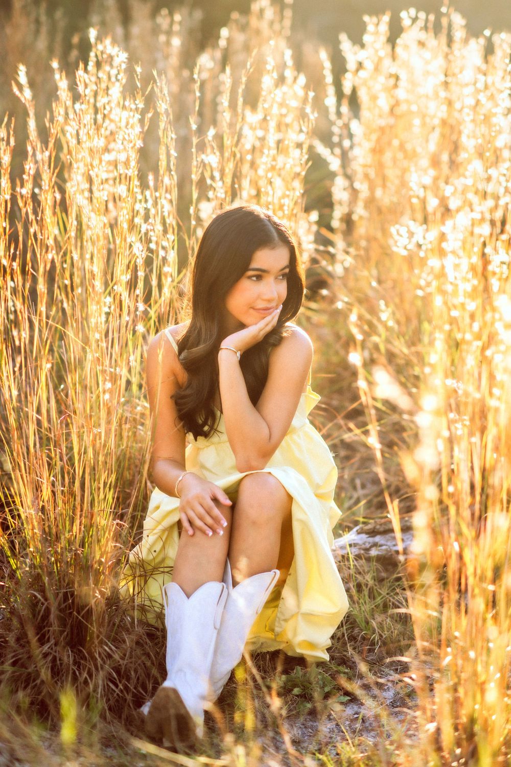 Young woman in yellow dress and white cowboy boots sitting in golden sunlit tall grass.