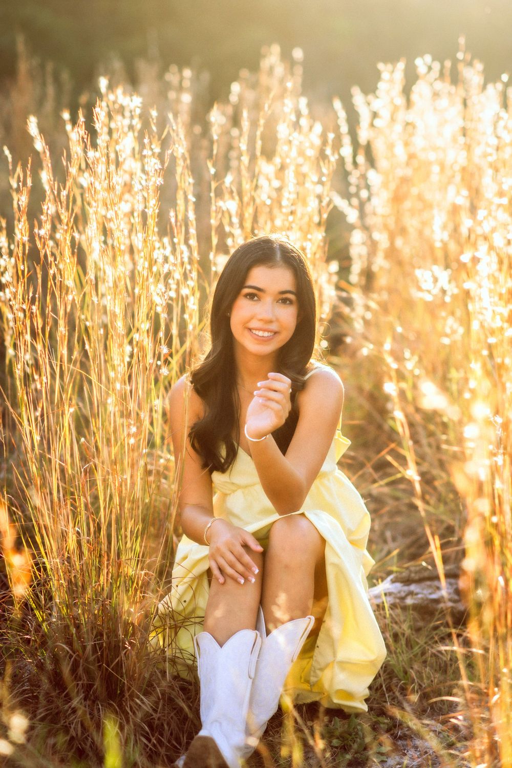 Smiling young woman in yellow dress and white cowboy boots sitting among tall golden grasses in sunlight.