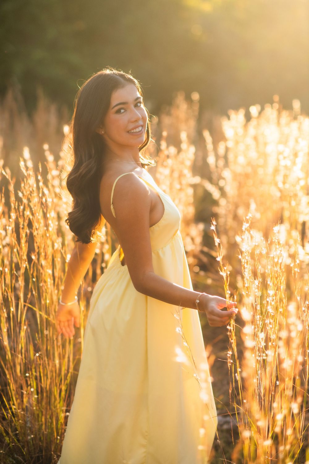 Brunette woman in yellow sundress standing among tall golden grasses in warm sunset light.