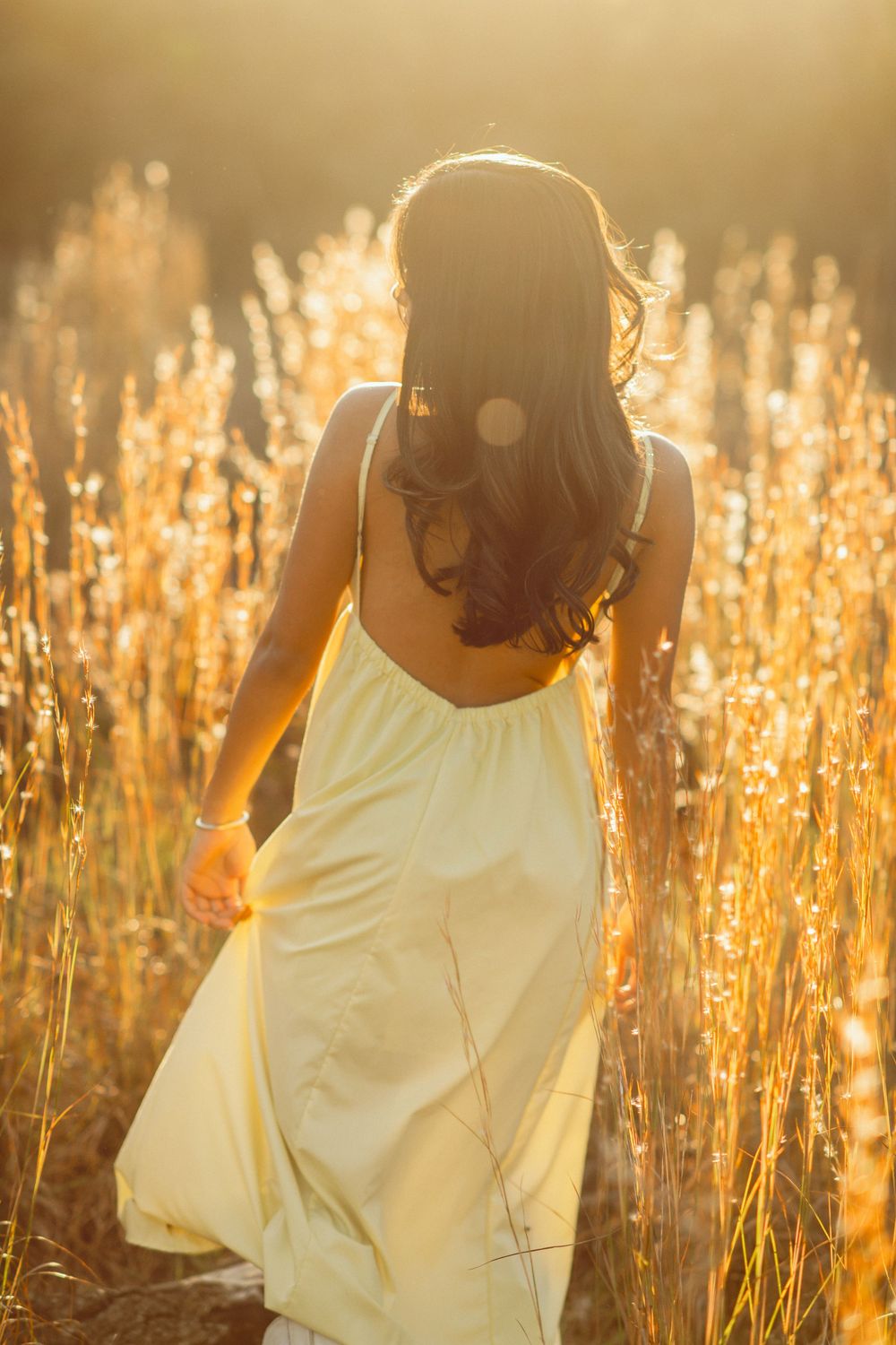 Back view of woman in yellow sundress walking through sun-drenched tall grass.