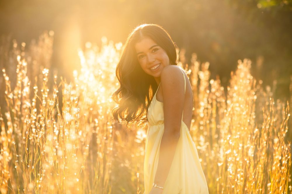 Smiling brunette woman in yellow dress surrounded by glowing golden sunlit tall grass.
