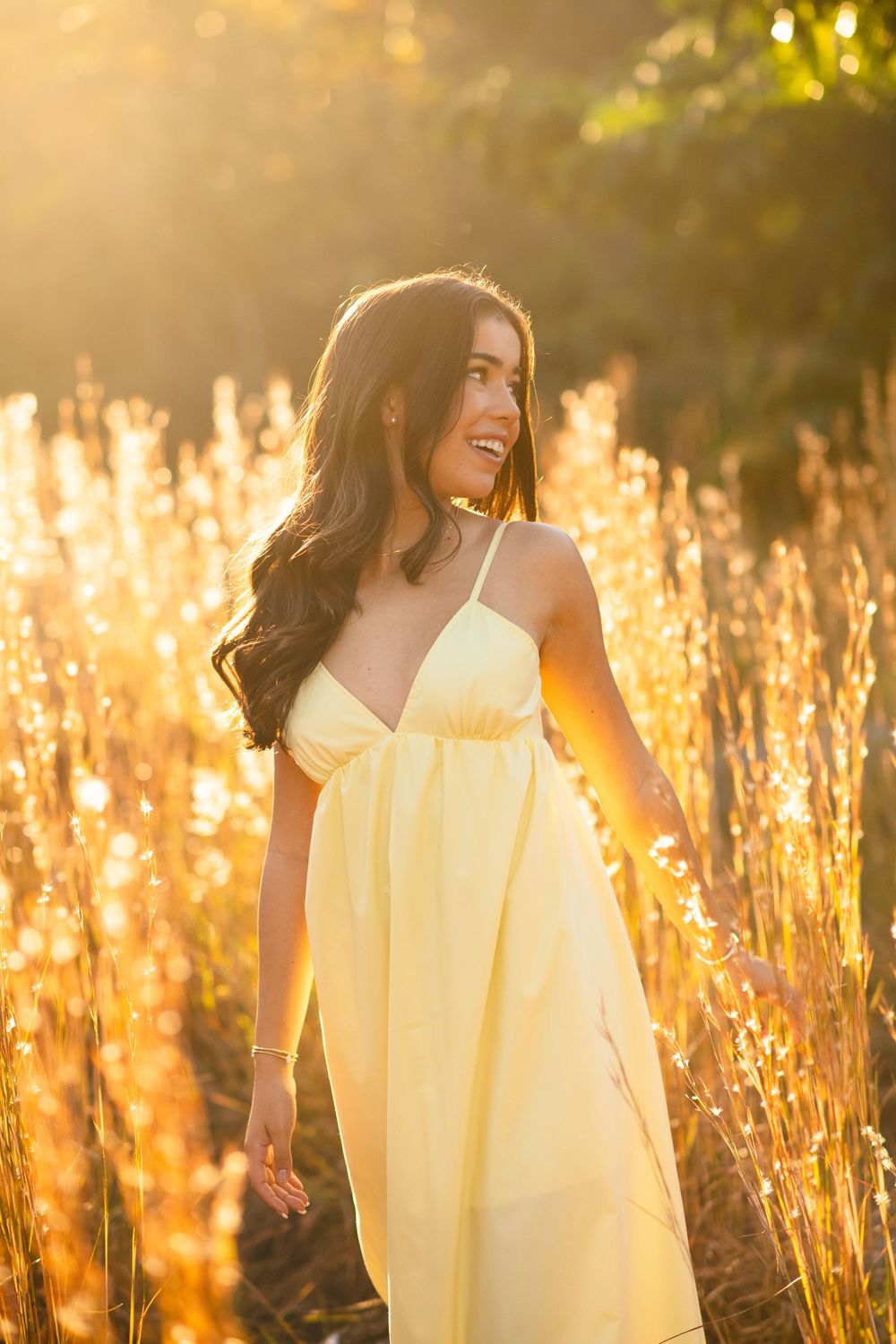 Laughing brunette woman in yellow maxi dress surrounded by glowing golden tall grass in bright sunlight.