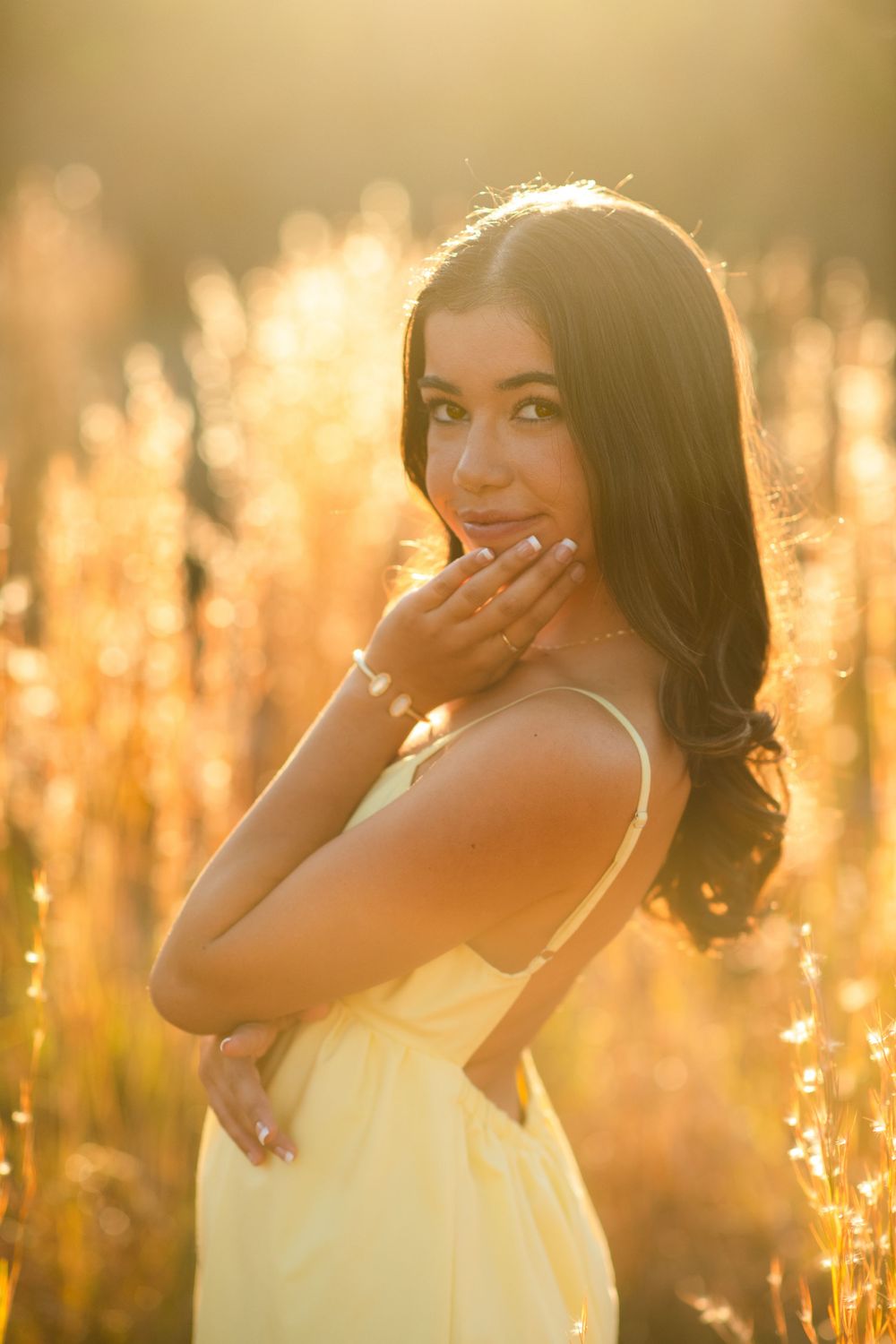 Young brunette woman in yellow sundress posing in glowing golden field at sunset.