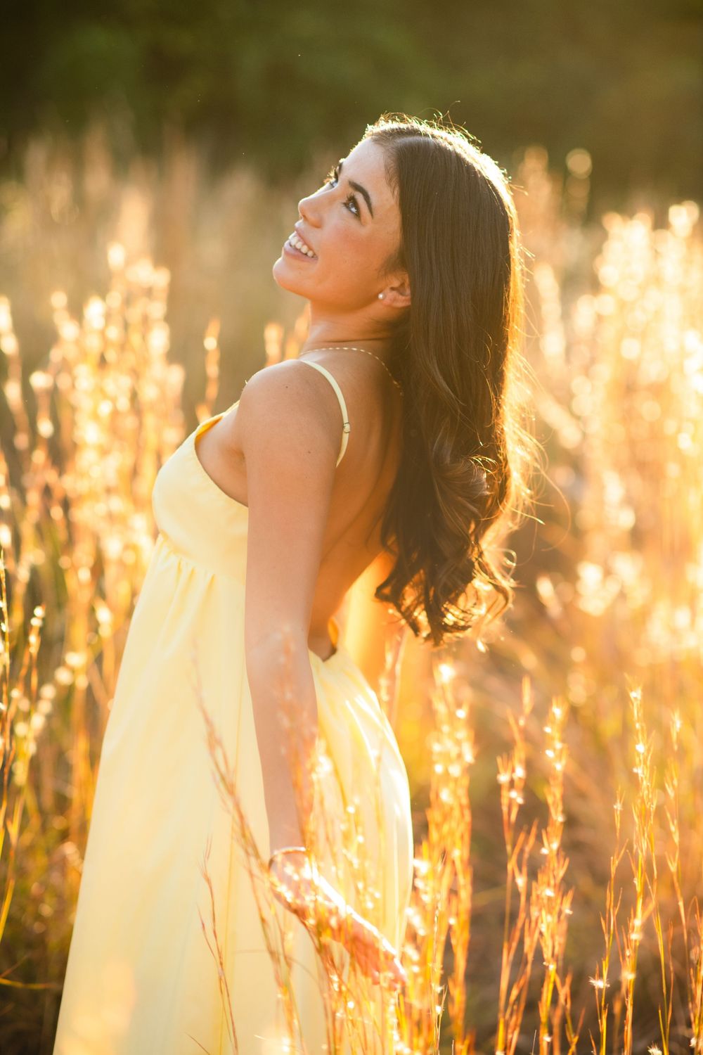 Smiling brunette woman in yellow sundress standing among tall grass in warm sunset light.