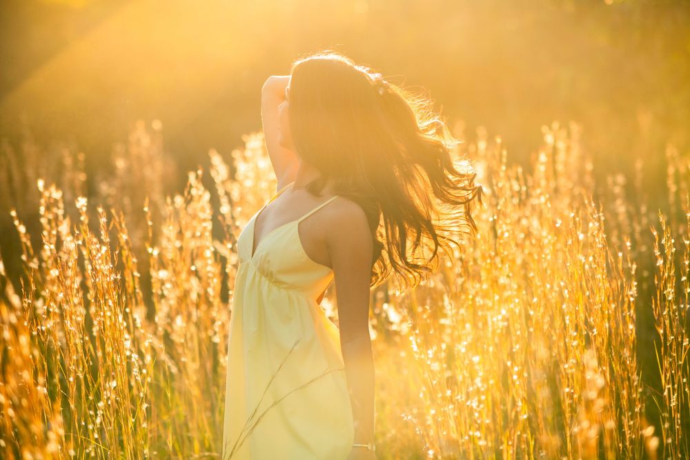 Silhouetted woman in yellow dress tossing hair in glowing golden sunset meadow light.