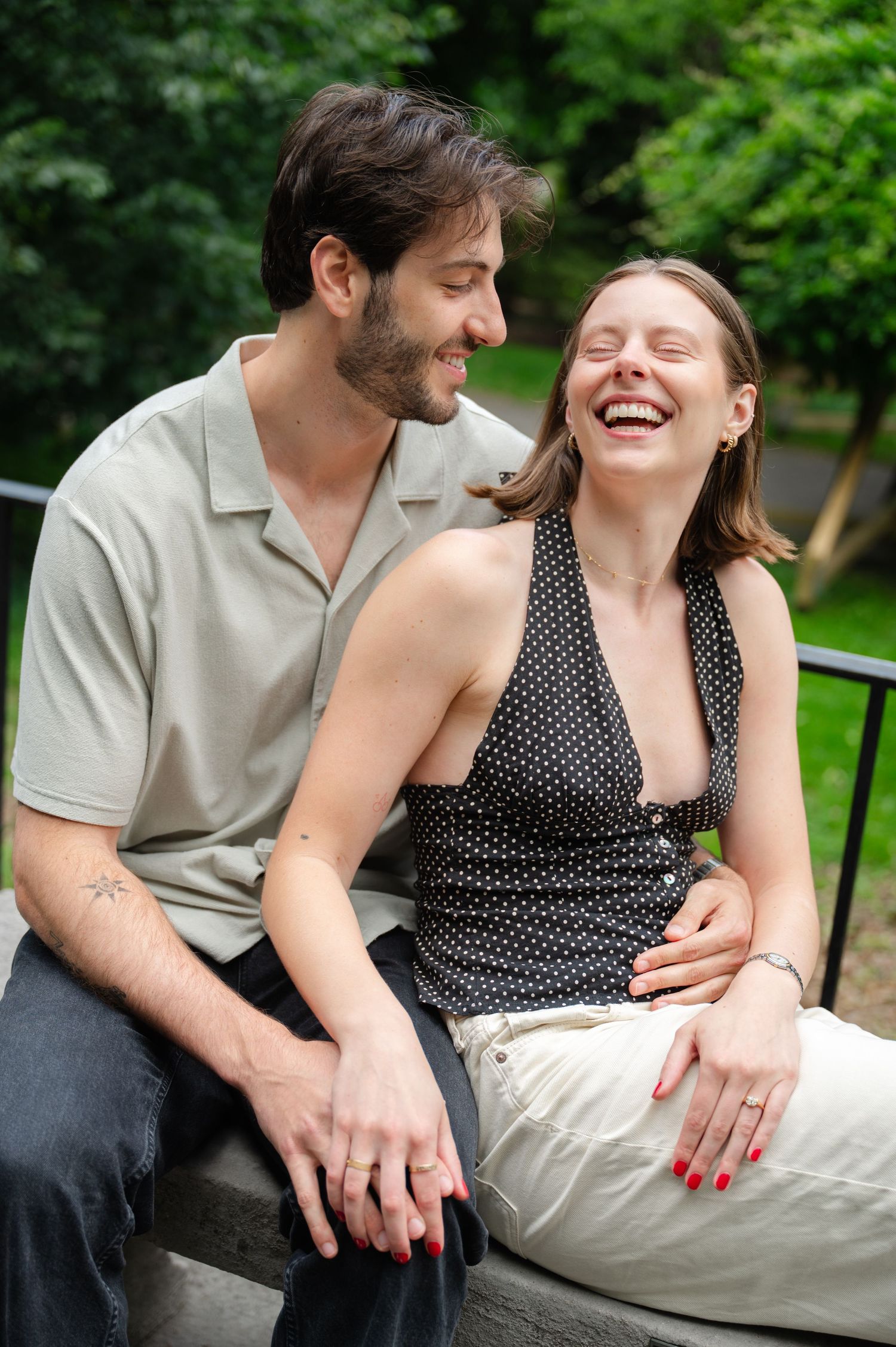 A laughing couple sits together on a bench in a lush green park, sharing a joyful and intimate moment.