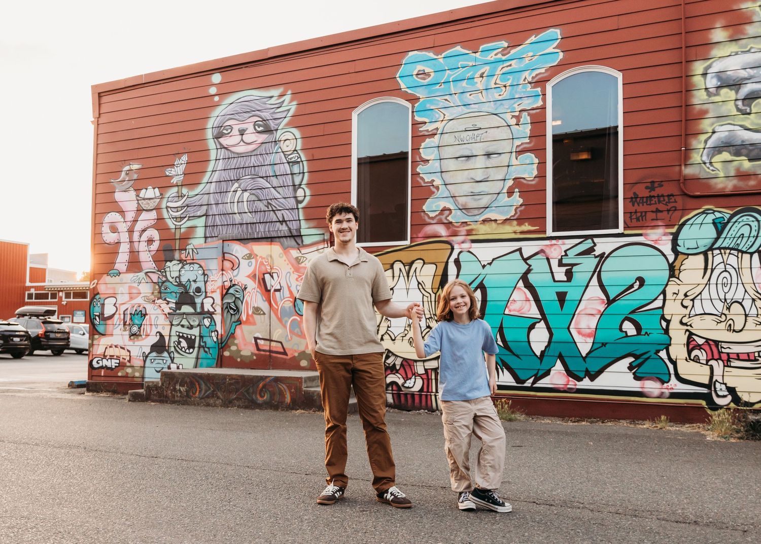 Two people pose in front of a colorful graffiti mural on a red building in an urban setting.