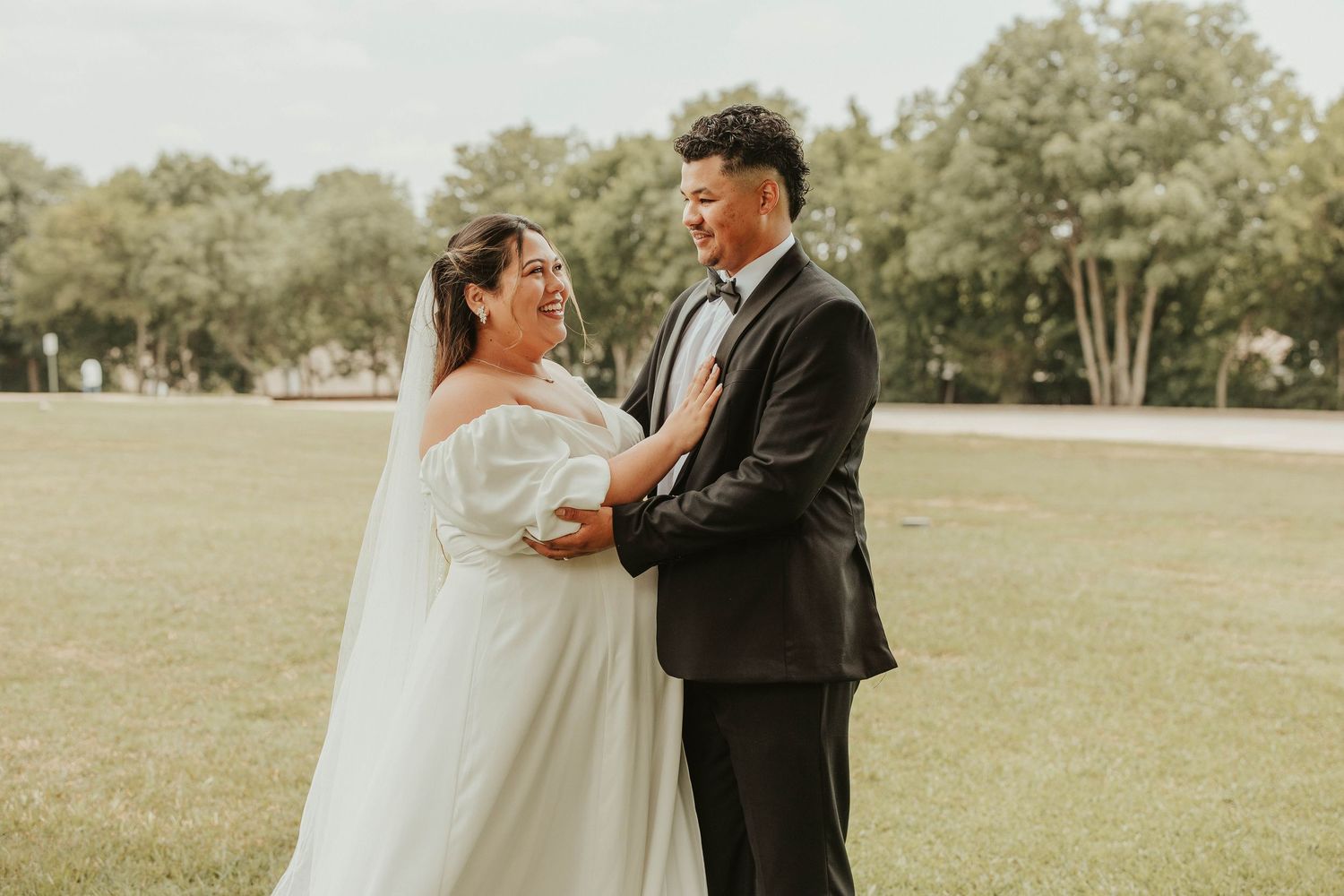 Bride in white off-shoulder gown and groom in black tuxedo share a joyful moment outdoors on green grass.