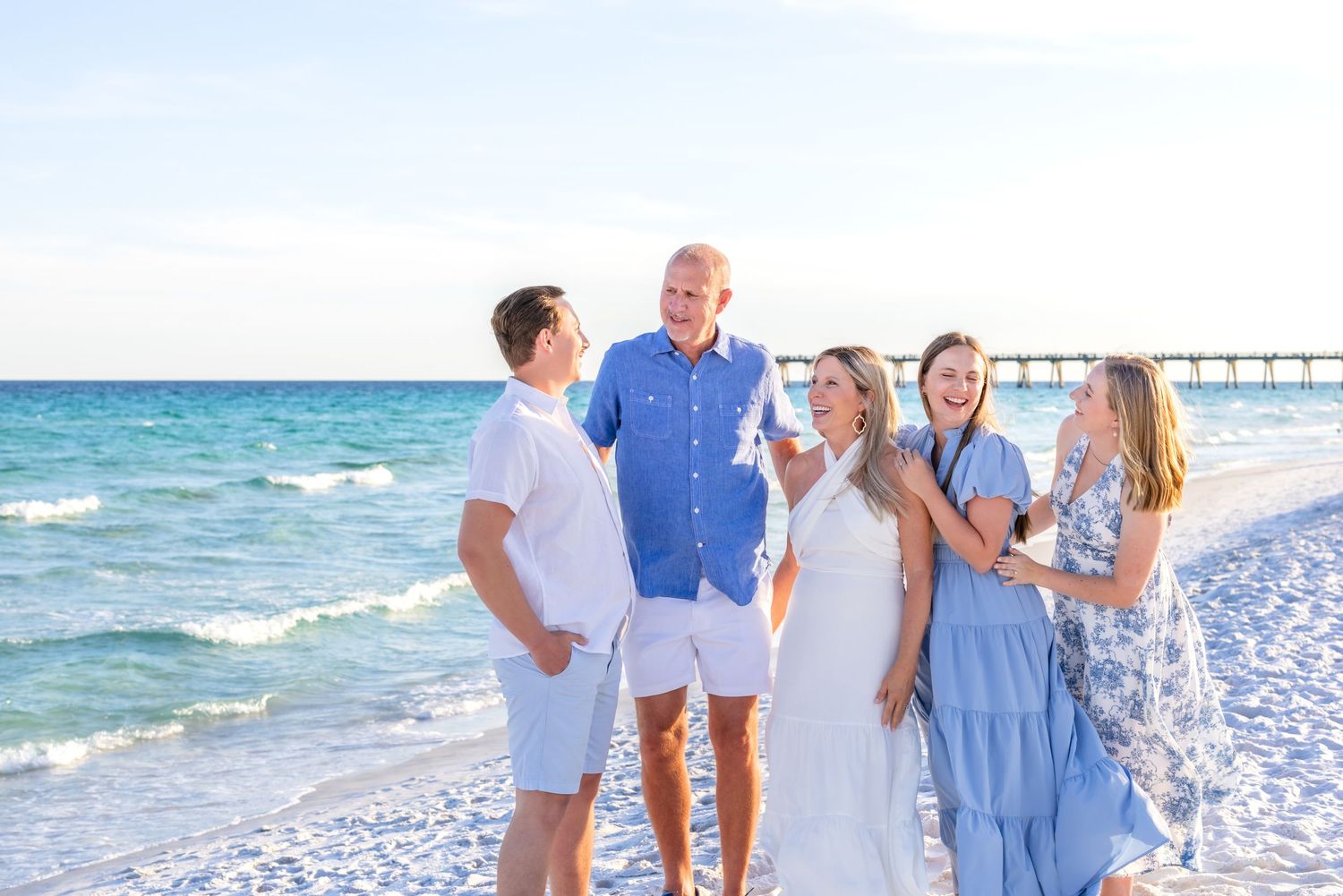 Family of five smiling together on a sunny beach with turquoise water and a pier in the background.