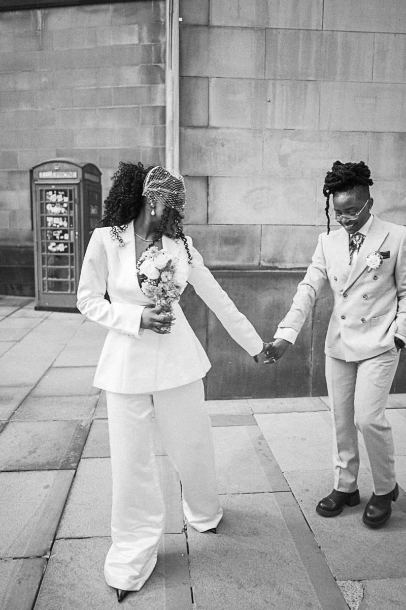 Black and white photo of two brides in white suits holding hands outside a stone building with a red phone box.