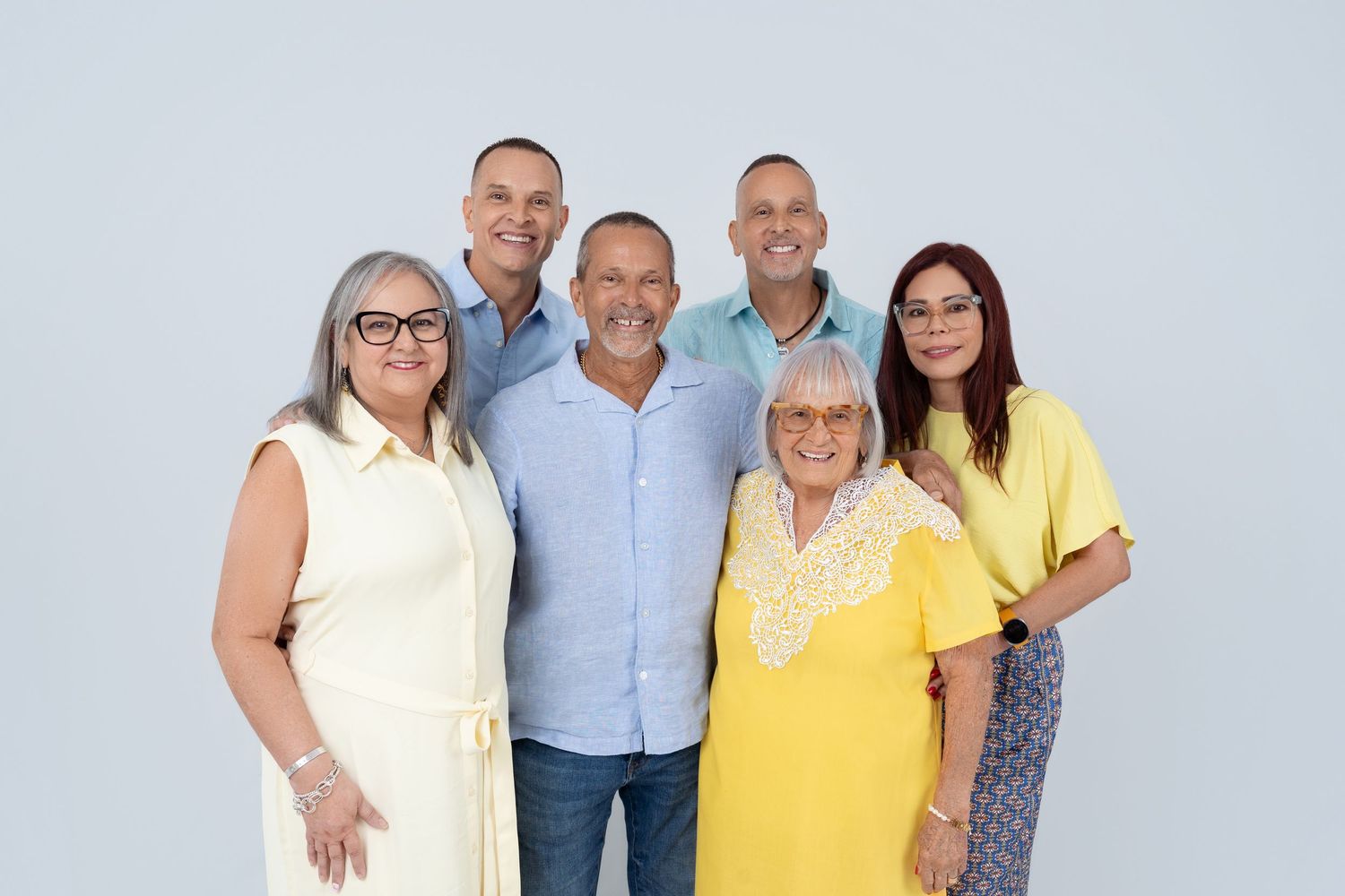 Six smiling adults pose together for a cheerful group portrait against a light gray background.