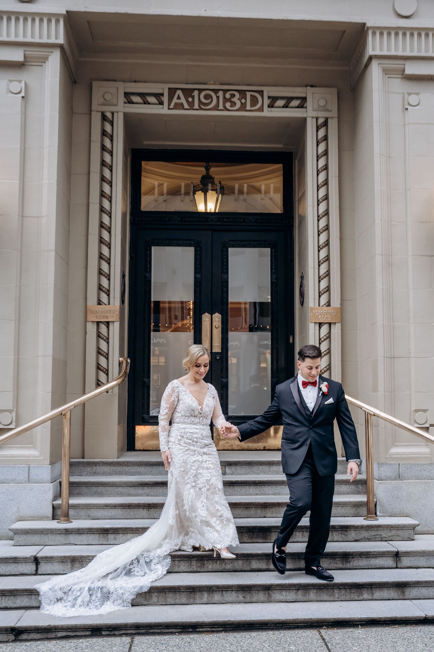 Bride and groom holding hands descending stone steps of a 1913 historic building, bride in lace gown with train.