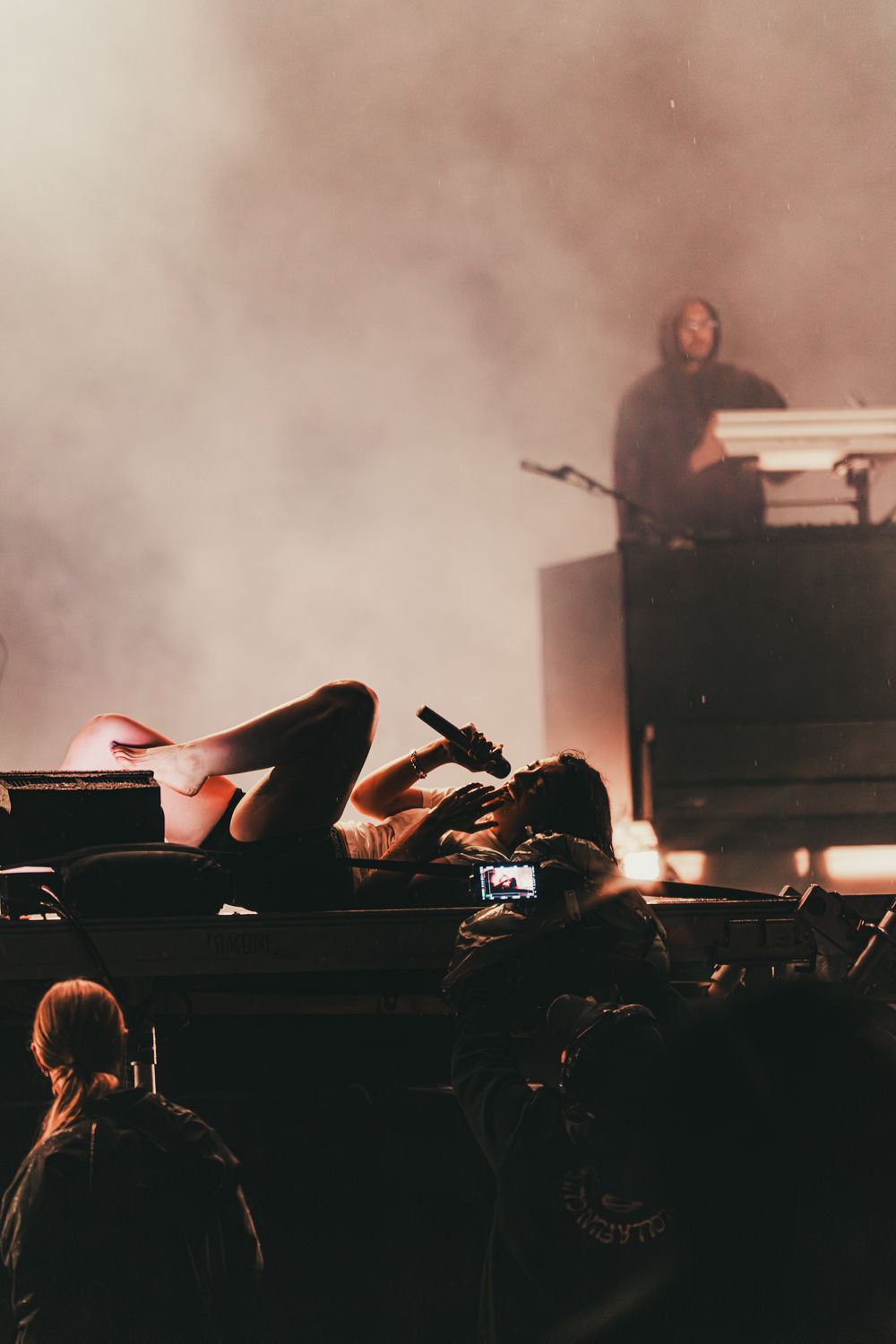 Performer lying on stage with microphone at smoky concert while crowd photographs and DJ performs in background.