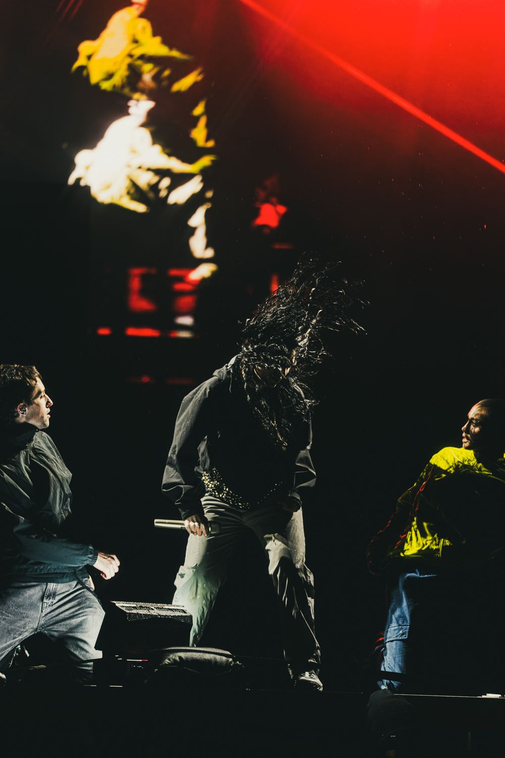 Three performers on a dark stage with red laser lights and a fiery projection backdrop during a live concert.
