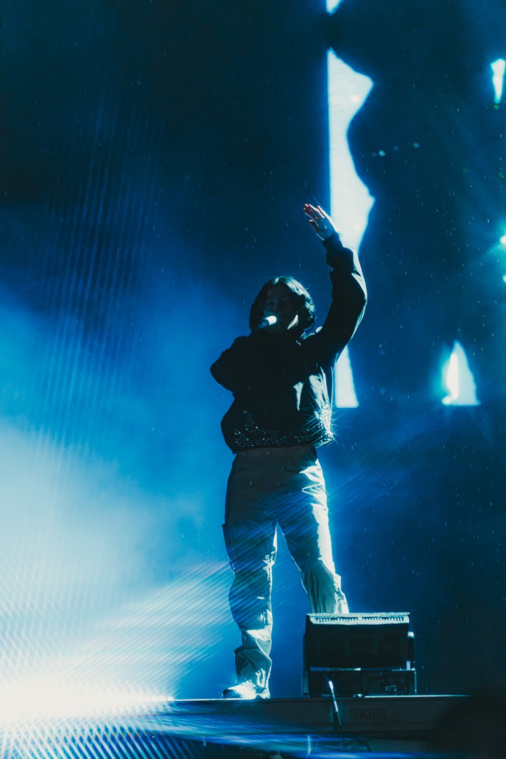 Performer on stage with arm raised, silhouetted against dramatic blue concert lighting and fog.